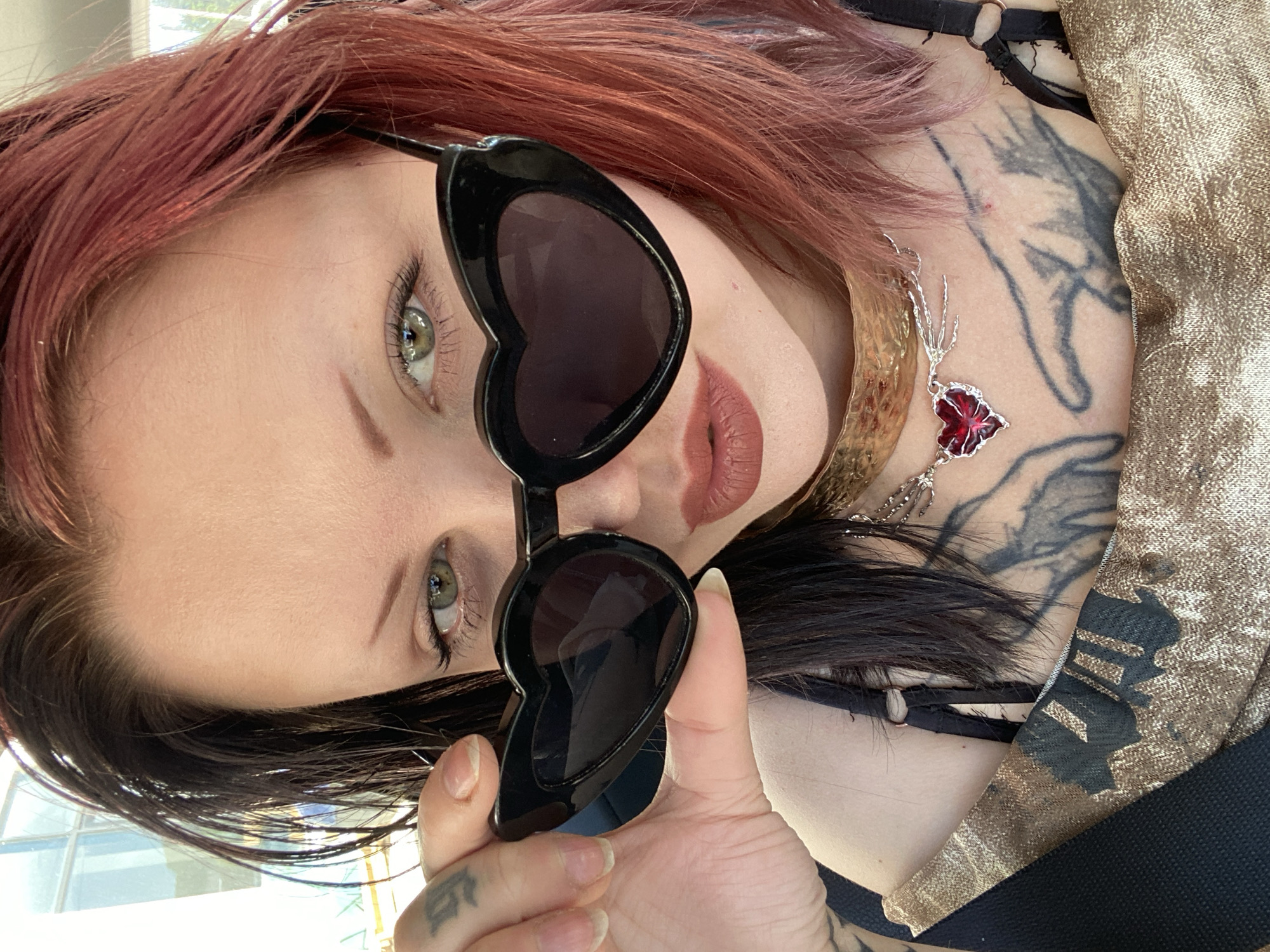 A woman with red and black hair is wearing heart-shaped sunglasses. She has a tattoo on her hand and another large tattoo on her neck and chest. She is also wearing a necklace with a red pendant. The background is blurry but appears to be outdoors with hints of sunlight. The location in Ulcinj, Montenegro cannot be determined.