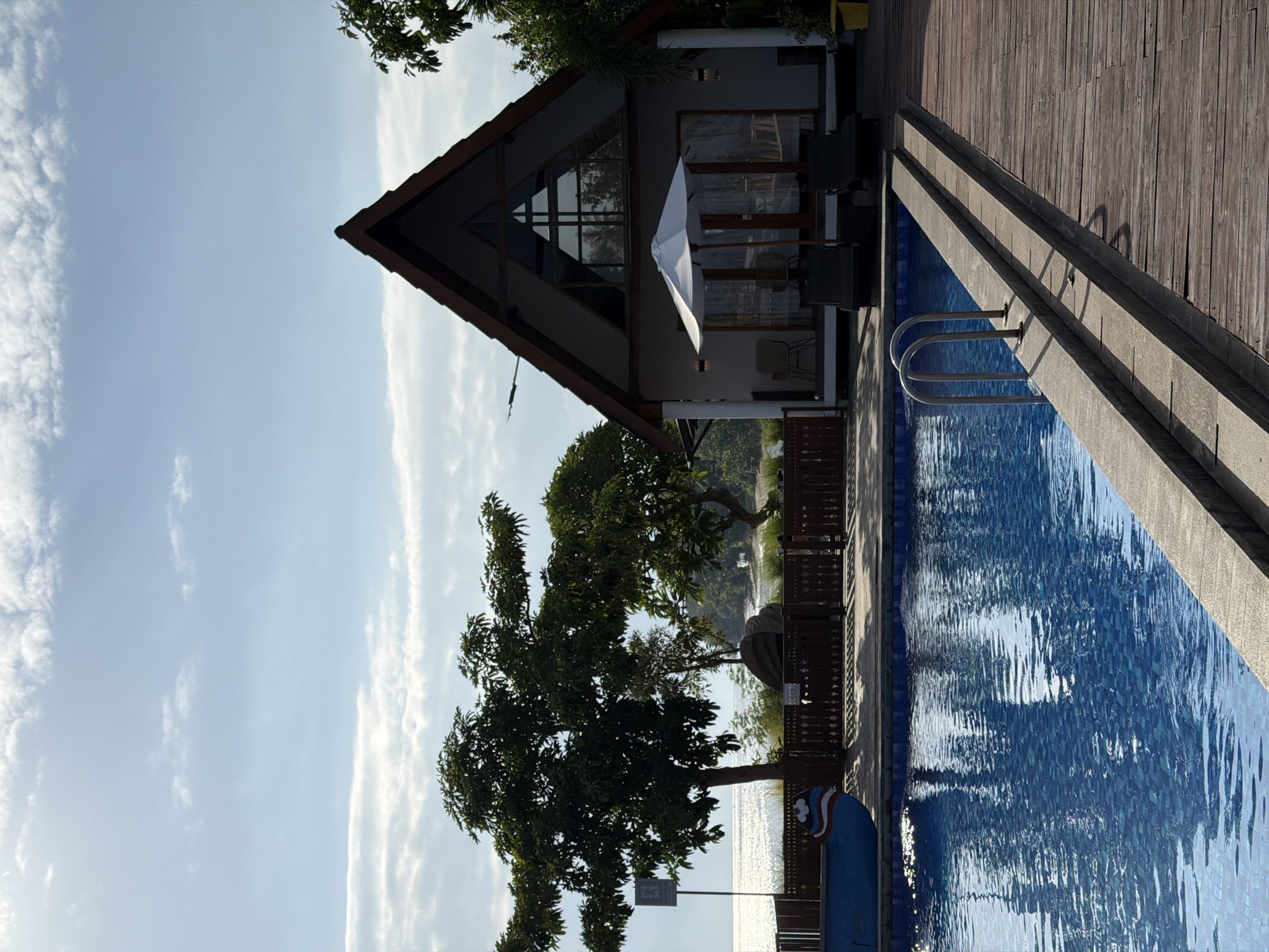 A resort setting is depicted in Pasir Putih, Indonesia, featuring a swimming pool and a triangular-roofed building. The pool, filled with blue water, has a tiled bottom and is bordered by a concrete ledge and a wooden deck. A metal ladder with two arched handrails provides access to the pool. In the foreground, a portion of a blue boat with red and white stripes is visible on the left. Behind the pool, a wooden fence separates the area from the building and surrounding vegetation.
The building, painted white, has a prominent triangular roof with dark brown trim. A large triangular window is situated at the peak of the roof. In front of the building, a white umbrella shades two dark lounge chairs. Two additional chairs and a small table are visible on the covered porch.
To the left of the building, large trees with dense green foliage are present, with the shimmering ocean and a distant shoreline visible in the background. The sky is a bright blue with scattered white clouds. The overall scene suggests a serene tropical environment.