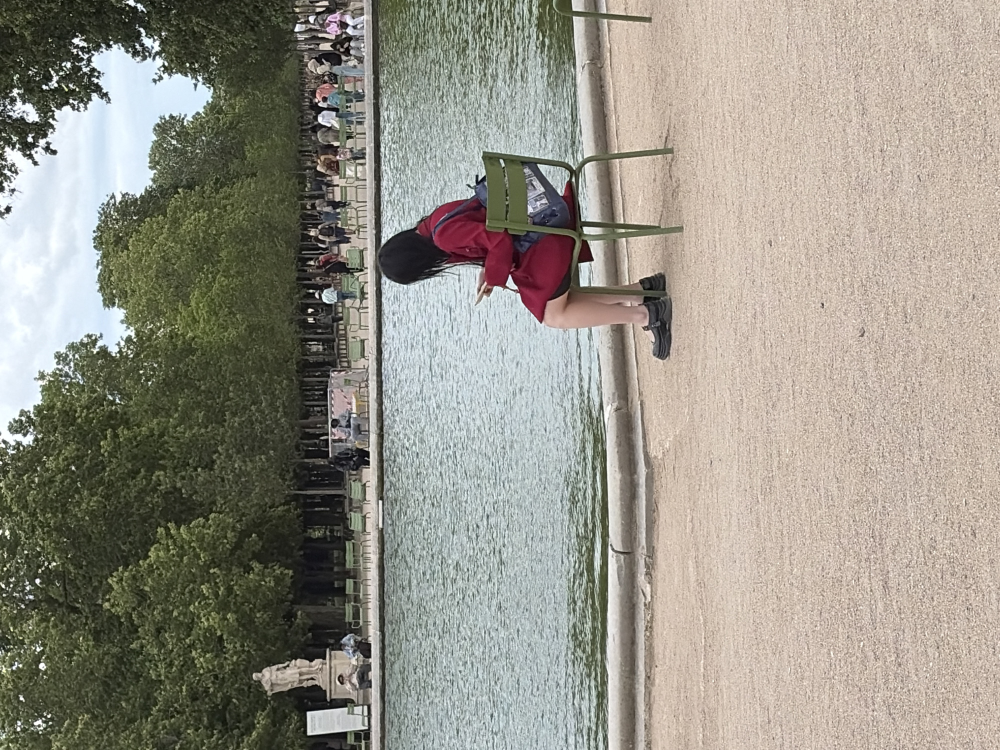 A young woman with dark hair, wearing a red coat, black skirt, and black platform shoes, sits alone on a green metal chair facing a large, calm body of water. She is holding a smartphone in her hands and appears to be looking at it. A blue backpack with a graphic print rests on the back of the chair. The scene is set in the Tuileries Garden in Paris, France, as indicated by the distinctive green chairs and the presence of a pond and statues typical of the park. In the background, there are rows of similar green chairs, trees, and a scattering of people walking and sitting, suggesting a leisurely afternoon. The ground in the foreground is covered with light-colored gravel. The sky is overcast.