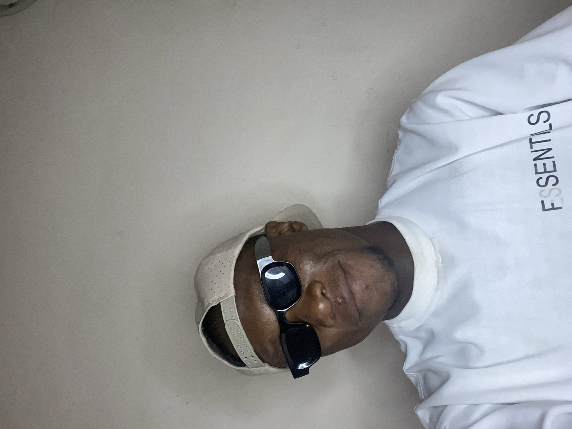 The image shows a man from the chest up, wearing a white "ESSENTILS" long-sleeved shirt, a light-colored baseball cap, and black sunglasses. He appears to be indoors against a light-colored wall. There is no indication of a location in Bukuru, Nigeria or any other location.