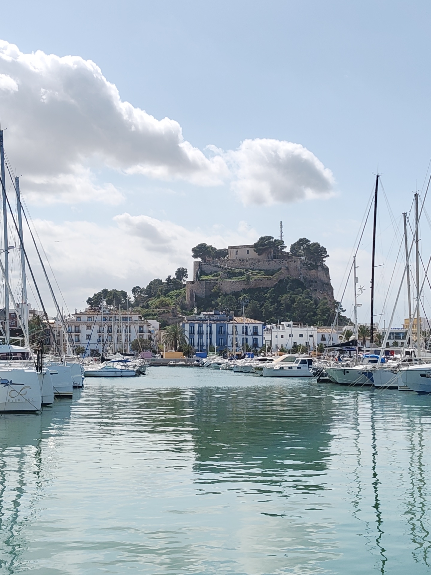 The image displays a panoramic view of the Dénia marina in Dénia, Spain, captured under a partly cloudy sky. The foreground is dominated by calm, light turquoise-green water exhibiting gentle ripples, reflecting boat masts and clouds. Multiple white sailboats are moored along both the left and right sides of the frame. On the left, the sterns of two vessels are visible, one labeled "EVERLAND" and another "BALI."
In the mid-ground, a strip of urban development lines the waterfront. This includes multi-story buildings, such as a white structure with visible text partially obscured but appearing to be "HOTEL" on its roofline, and a prominent blue and white building to the right identified as "HOTEL EURASET." Palm trees are interspersed among these structures.
The background is defined by a steep, green, tree-covered hill, atop which sits the historic stone complex of Dénia Castle. Features of the castle, including walls and battlements, are discernible. A modern cellular communication tower is positioned on the hilltop near the castle structures. The sky above is a light blue with scattered white cumulus clouds. No individuals or specific actions are visible within the frame.