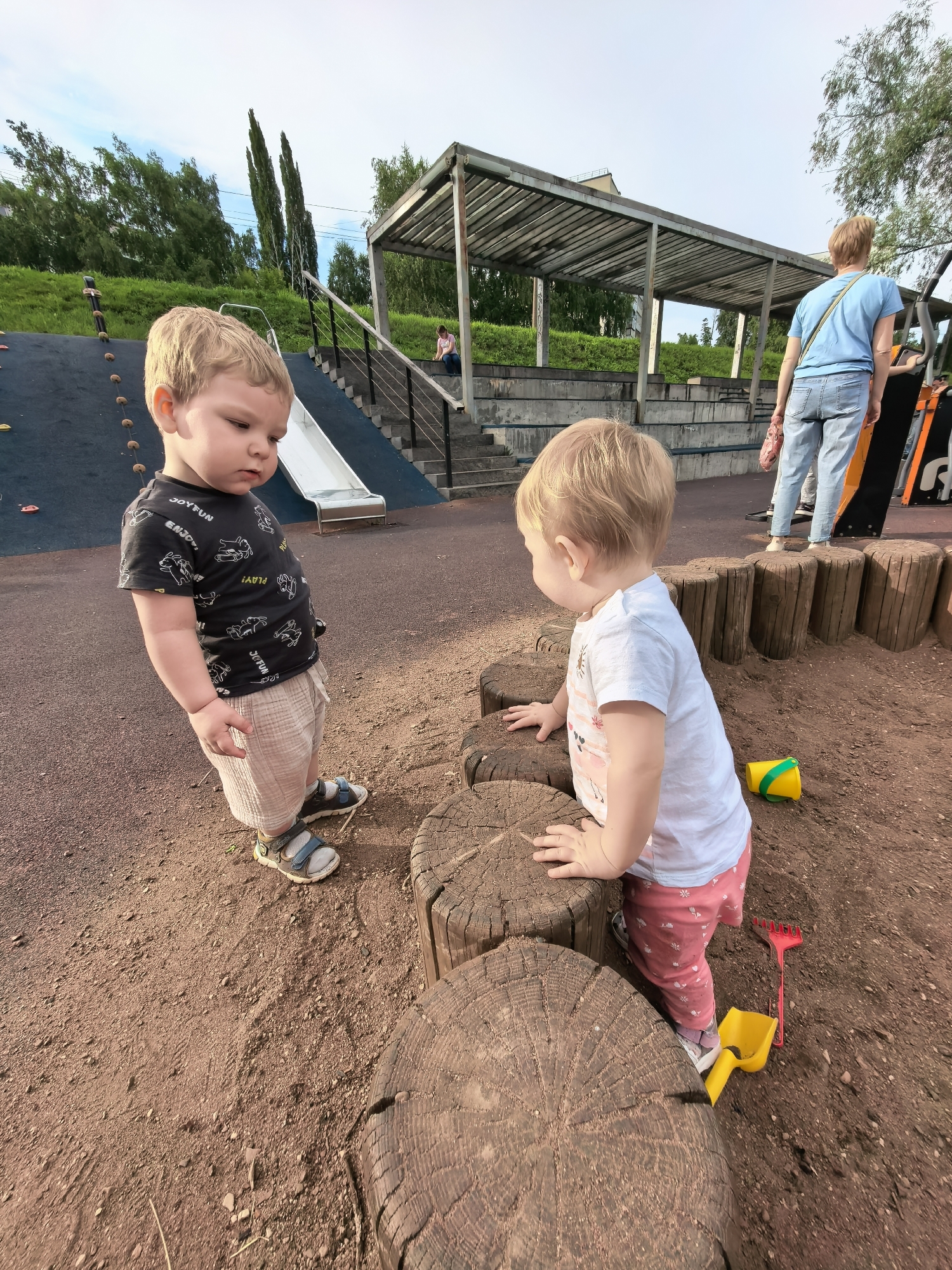 This is a medium shot of two young children, a boy and a girl, at a playground. The boy, facing left, has blond hair and is wearing a black t-shirt with a white graphic print and light-colored shorts. He is looking down with a serious expression. The girl, facing right, also has blond hair and is wearing a white t-shirt with a pink graphic print and pink pants with a floral pattern. She is reaching out with her hands towards some wooden logs in the foreground.
The playground has a sandy, reddish-brown surface. The wooden logs appear to be part of a climbing or balancing structure. In the background, there is a blue slide with a silver chute, stairs leading up to it, and a covered seating area or pavilion with a metal roof. A woman in a blue t-shirt and jeans is visible further in the background, near the pavilion. There are also children's toys on the ground, including a yellow bucket and a red rake.
The time of day appears to be late afternoon or early evening, given the lighting. The sky is partly cloudy. The overall mood is one of playful exploration. The setting is an outdoor playground, likely in a park.
The text on the boy's t-shirt is partially visible and appears to say "JOY FUN" and "ENJOY".