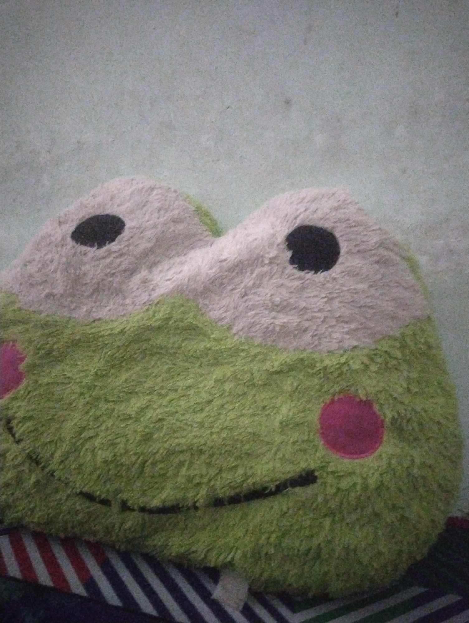 The image shows a close-up view of a green stuffed frog toy with a white face, black eyes, and pink cheeks. The toy is fluffy and appears to be resting on a striped surface. The background is a pale green, textured wall. No other objects or people are visible. The image is indoors and well-lit.
