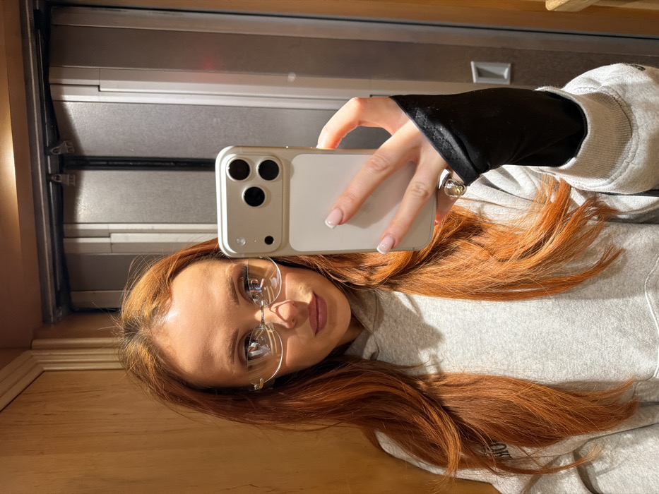 A young adult female with long, reddish-orange hair and fair complexion takes a mirror selfie in an enclosed space in the city of Paris, France. She wears rimless eyeglasses with silver-toned frames and a light grey hooded sweatshirt. A black long-sleeved garment is visible beneath the cuff of her right sleeve. Her right hand, featuring a French tip manicure and a silver-toned ring on the ring finger, holds a light-colored smartphone, likely an iPhone, equipped with a triple camera system and a matching case. The background consists of light-toned wood paneling on the left and a two-panel, silver-grey metal door with visible latch mechanisms at the top center, consistent with an elevator.