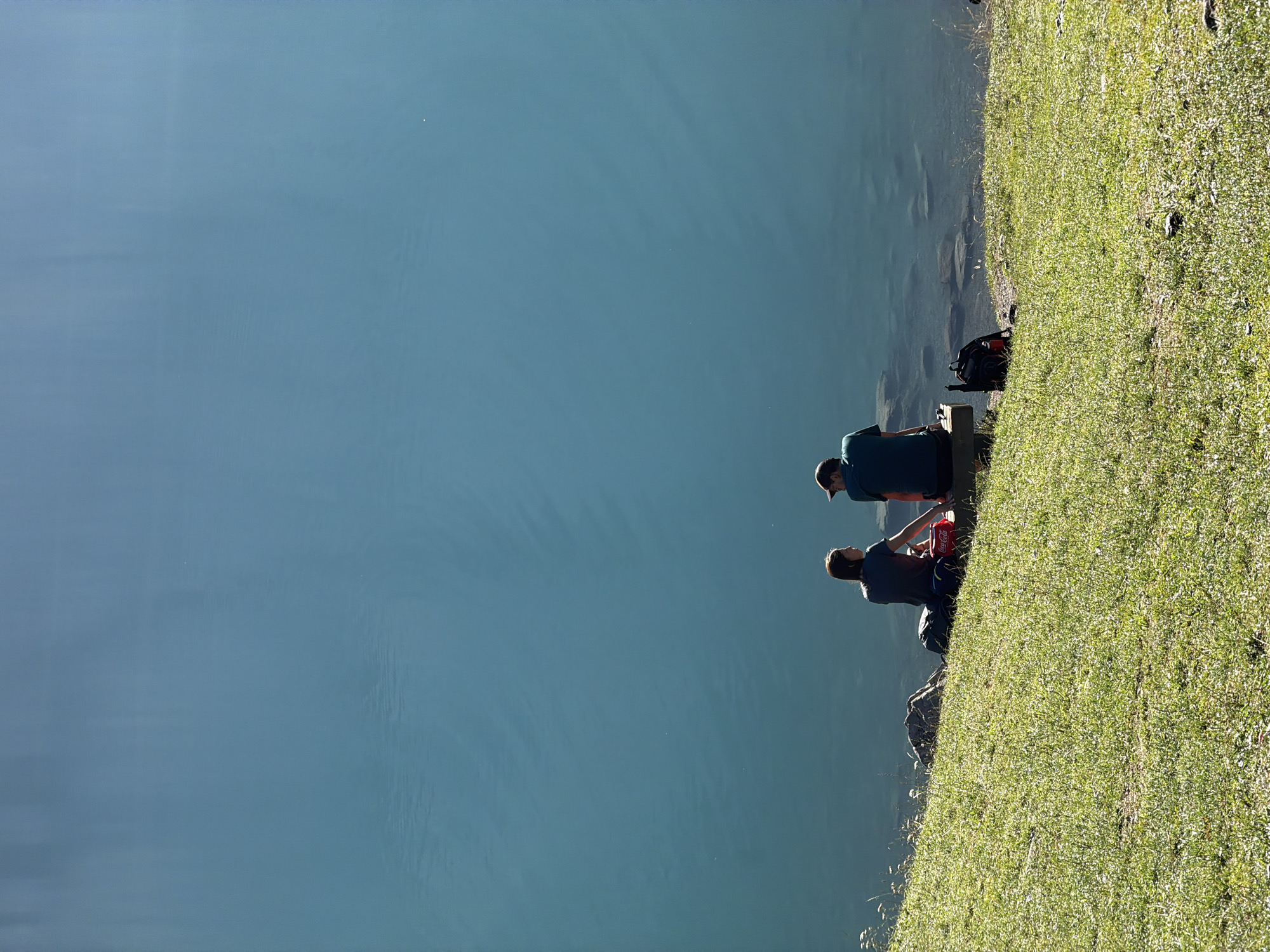 Two individuals, one female and one male, are seated on a bench at the edge of a tranquil, expansive body of water. The female is wearing a dark t-shirt and jeans, while the male is dressed in a dark t-shirt and a baseball cap. Both are facing away from the camera, gazing out at the water. The setting appears to be a natural landscape, with a grassy embankment sloping down to the water's edge. Several backpacks and a cooler bag are placed on the ground near the bench. The water itself is a deep, clear blue, reflecting the sky above. The lighting suggests it is daytime, with the sun casting shadows on the grass. This scene is located at the Oeschinensee lake near Kandersteg, Switzerland, a renowned alpine lake known for its stunning turquoise waters and dramatic mountain surroundings.