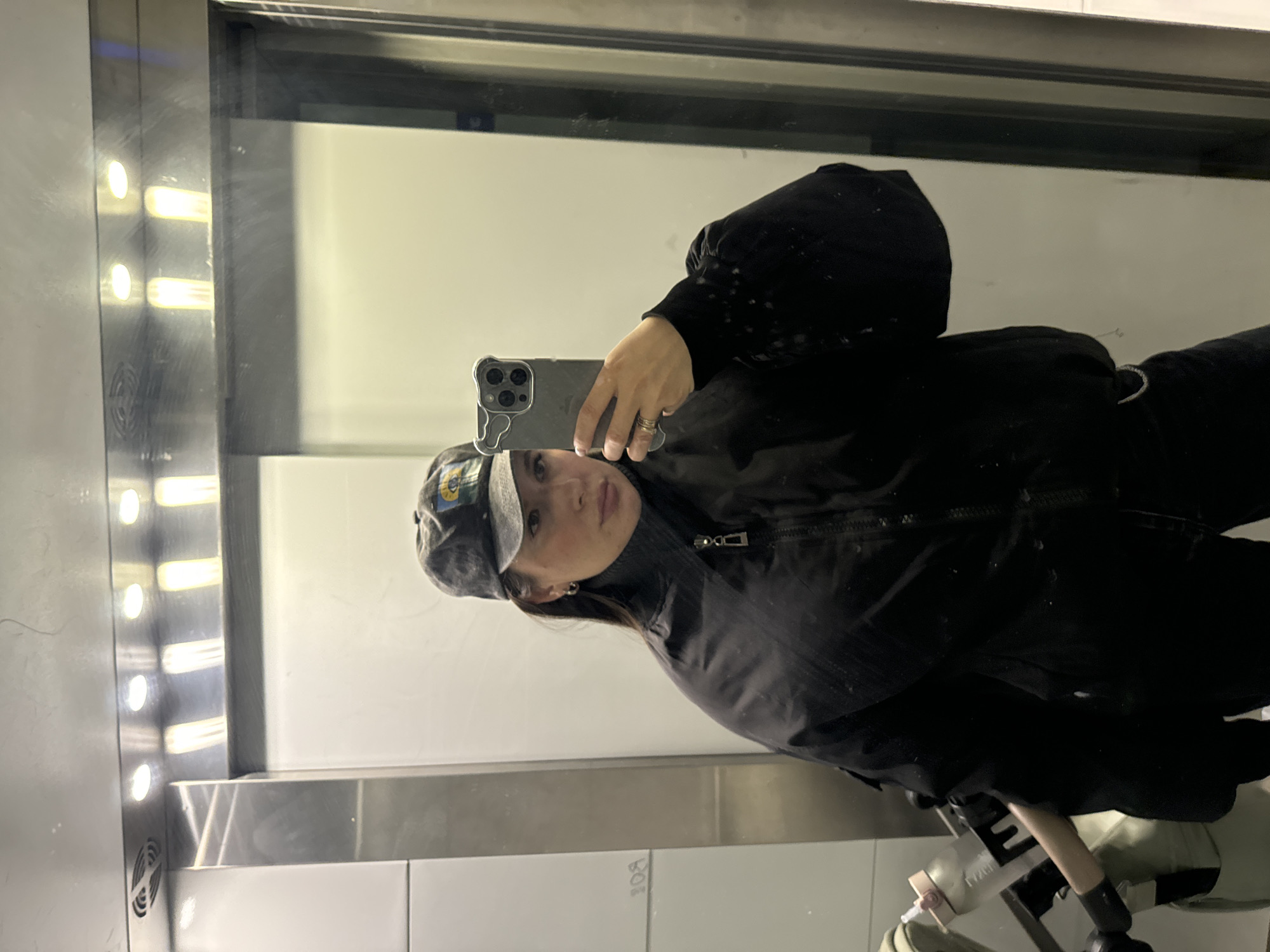 A person is taking a selfie in an elevator. The person is wearing a black jacket, a black turtleneck, and a gray baseball cap. The person is holding a smartphone in their left hand. The elevator has a mirrored wall and metal accents. The location is Селекционер-2 Снт, Russia.