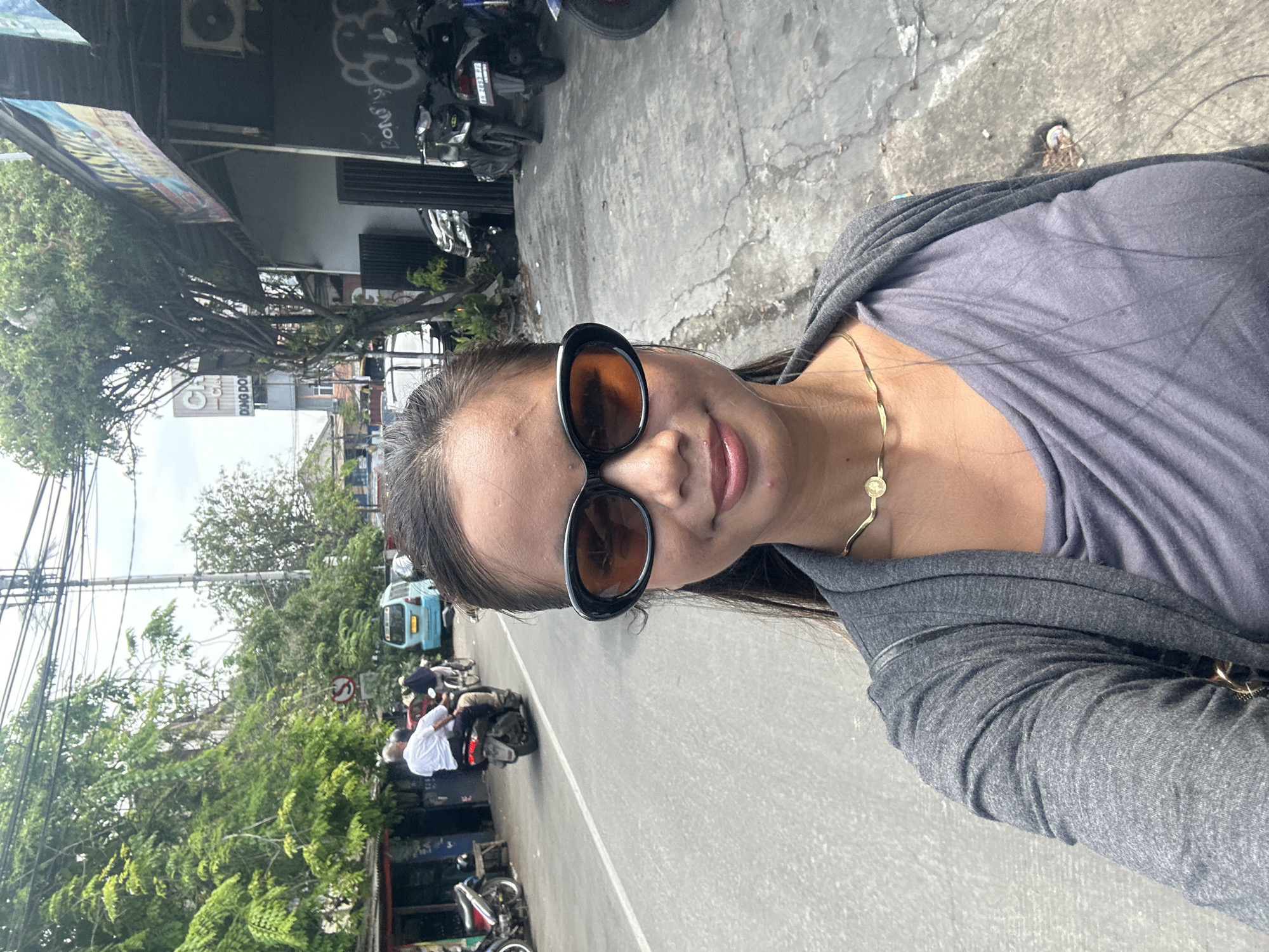 A woman with long dark hair, wearing sunglasses, a gray top, and a gold necklace, takes a selfie on a city street. Behind her is a street with motorbikes parked along the curb and moving along the road. Across the street, trees line the sidewalk, and buildings are visible in the background. There are overhead wires and signs on the buildings. Based on the street signs and the style of buildings, the location appears to be in North Jakarta, Indonesia.
