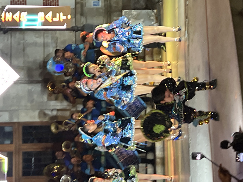 The image captures an outdoor nighttime cultural performance, likely in a public square or street in the city of Arica, Chile. In the foreground, a male dancer is positioned centrally, bent low with flexed knees, his head bowed to the right. He wears an elaborate dark costume with green accents, metallic embellishments on his limbs, and a large, dark, possibly feathered or tasseled headpiece. To the immediate left, a hand holds a smartphone on a selfie stick, pointed towards the performers.
Behind the male dancer, approximately eight individuals, primarily female, are dancing. They are uniformly dressed in elaborate, brightly colored costumes, predominantly blue with silver sequins, fringe, short skirts, and decorative headpieces. Some wear high-heeled shoes. One female dancer on the left holds a cylindrical drum. Another female dancer in the center-right is depicted with her mouth open, mid-action, holding a small, indistinct object.
In the background, behind the dancers, a musical ensemble consisting of approximately 10 to 15 individuals, predominantly male, performs as a brass band. They play various instruments including at least three tubas or sousaphones, trumpets, and trombones, and wear matching light blue shirts. The backdrop is a multi-story, light-colored stone building featuring large, dark wooden double doors. To the right of this building, a vertical neon sign, partially obscured, displays the word "ZARBALT." The scene is illuminated by strong artificial stage lighting, highlighting the performers against the dark nighttime environment. The ground surface appears to be paved.