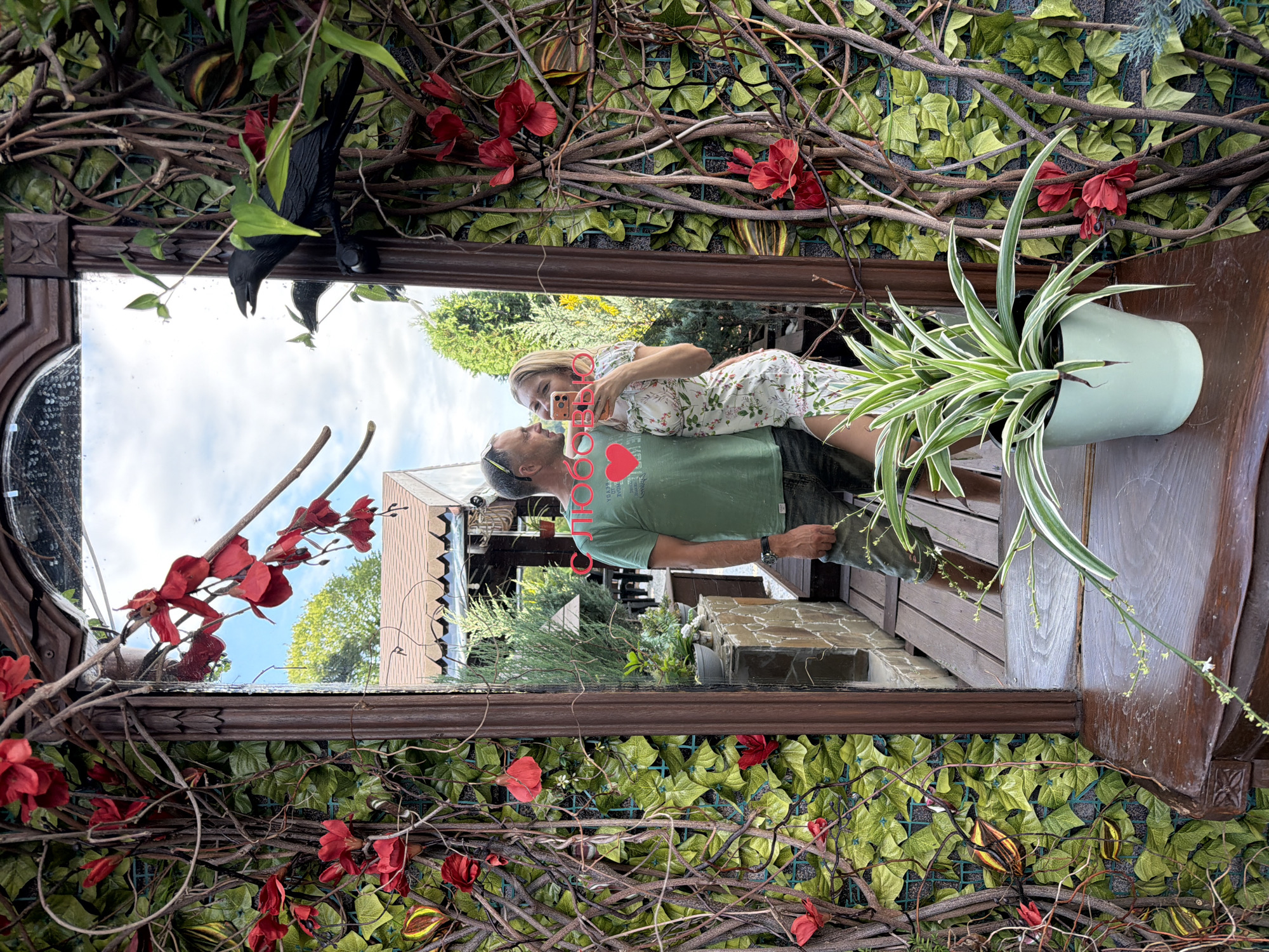 A vertical, reflected medium shot shows a couple kissing in front of a mirror adorned with artificial ivy and red flowers. The mirror is framed by a dark, ornate wooden border.
The couple, a man and a woman, are reflected in the mirror. The woman, with blonde hair, is wearing a floral dress and holding a pink smartphone, appearing to take a selfie. The man, wearing a green t-shirt with a red heart graphic and a black watch, is looking at her. Their faces are close, suggesting an intimate moment. The red heart graphic on his shirt contains white text that is not fully legible.
Behind the couple, the reflection shows an outdoor setting with trees, a wooden structure, and a blue sky with some clouds. The overall impression is of a warm, pleasant day. The scene is set outdoors, possibly in a garden or patio area, given the presence of plants and wooden furniture.
In the foreground, to the right of the mirror, a spider plant in a pale mint green pot is visible. The artificial ivy and red flowers surrounding the mirror suggest a decorative arrangement, perhaps at a cafe or an outdoor event. A black crow-shaped ornament is also visible at the top left of the mirror frame, adding a whimsical touch.
The image is taken at an angle, with the reflection appearing upside down relative to the viewer's perspective. The time of day appears to be daytime, indicated by the bright sky and shadows. The location context is Nizhnyaya Shilovka, Russia.