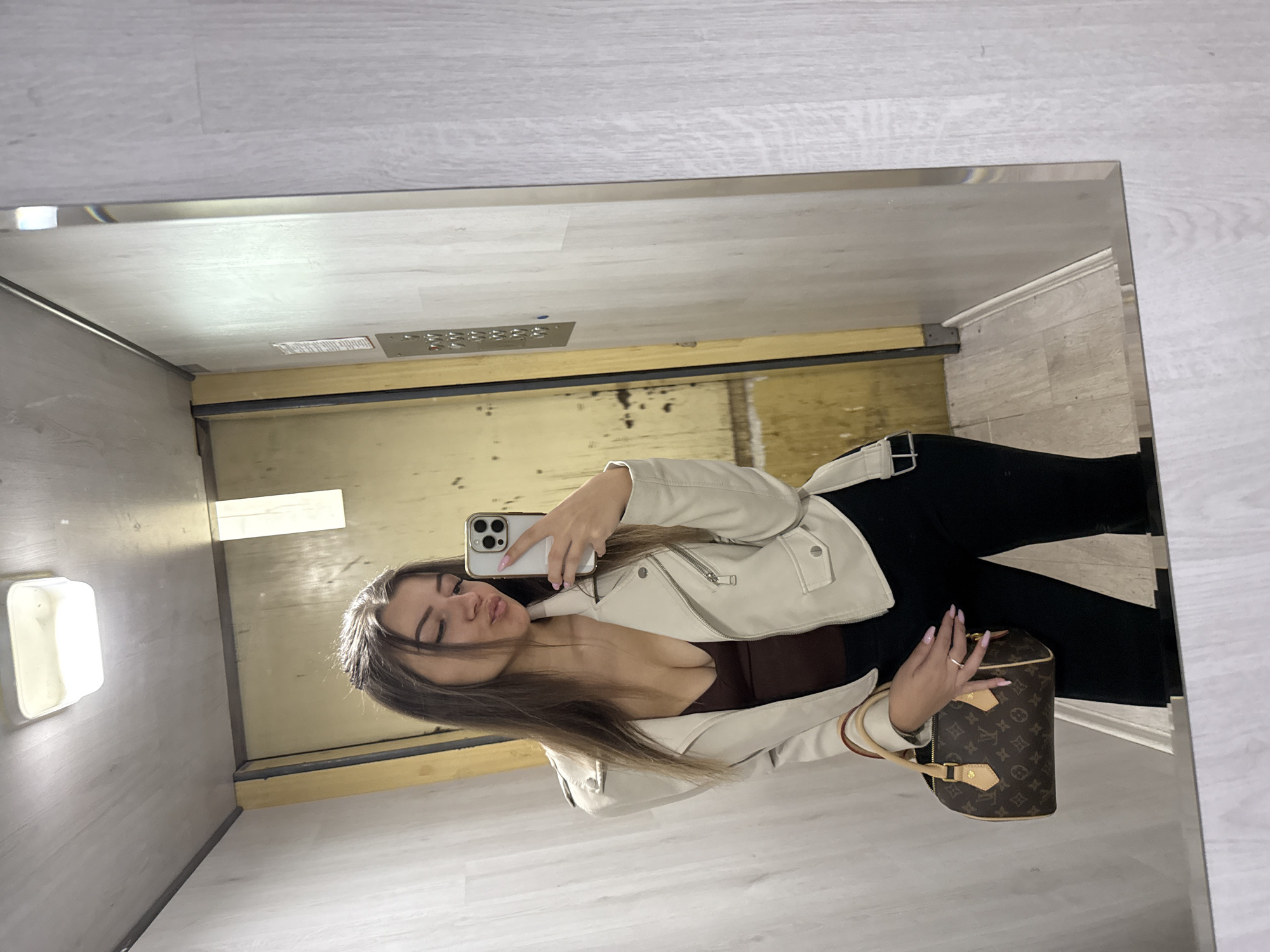 A woman is taking a mirror selfie inside an elevator. The elevator's interior is utilitarian, featuring light-colored walls and basic lighting. The woman is wearing a light-colored jacket over a dark top, paired with black pants. She is holding a Louis Vuitton handbag. The elevator appears to be in Sofia, Bulgaria, although there is no specific landmark to pinpoint the exact location.