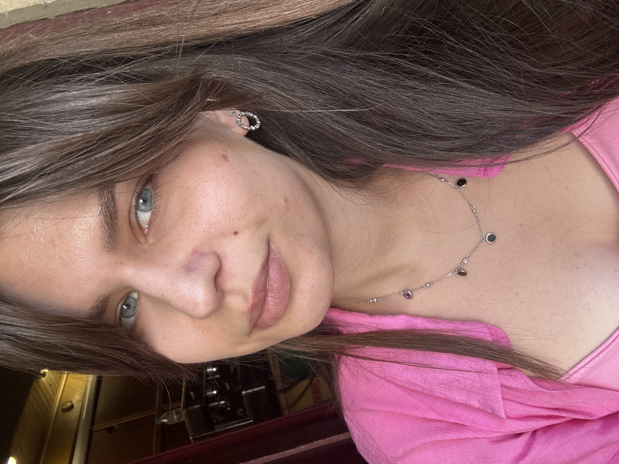 In an indoor setting, a young Caucasian woman with blue eyes and brown hair is captured in a close-up, shoulder-up portrait. She is wearing a bright pink shirt and a silver necklace with small circular pendants. Her hair is dark brown and styled with bangs framing her face. The background is slightly blurred, suggesting an indoor environment, possibly a cafe or a shop, with hints of wooden elements and a dark object that might be a piece of equipment. The lighting is natural, illuminating her face and upper body. She is looking directly at the camera, with a neutral expression.