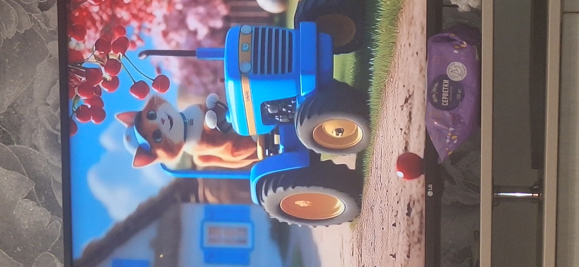 An animated orange and white cat wearing a blue cap and collar is driving a blue toy tractor with yellow wheels. The cat is on a dirt path in front of a house with blue shutters. In the background are pink trees with red cherries. The scene is being viewed on a television screen, which is placed on a stand. A purple package of wet wipes is on the stand in front of the television. The LG logo is visible on the bottom bezel of the television. The image does not contain any information about Bila Tserkva, Ukraine.
