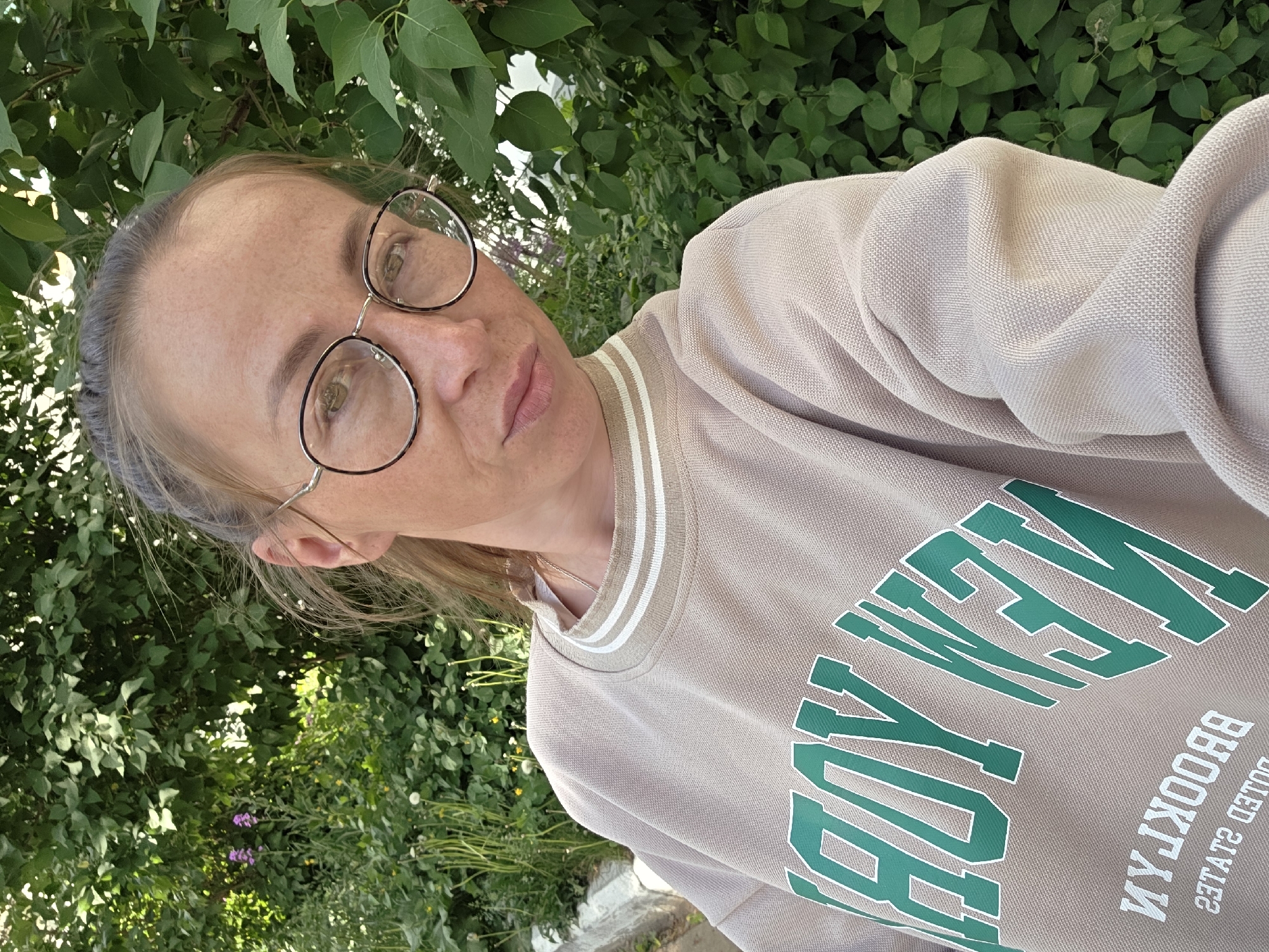 A woman is posing for a selfie. She has light skin, freckles, and brown hair pulled back in a ponytail. She wears round glasses and a beige sweatshirt with green lettering that says "New York" and "Brooklyn." The background is filled with lush green foliage, suggesting an outdoor setting, possibly a garden or park. The image's location and its connection to Chaikovsky, Russia, are not discernible.