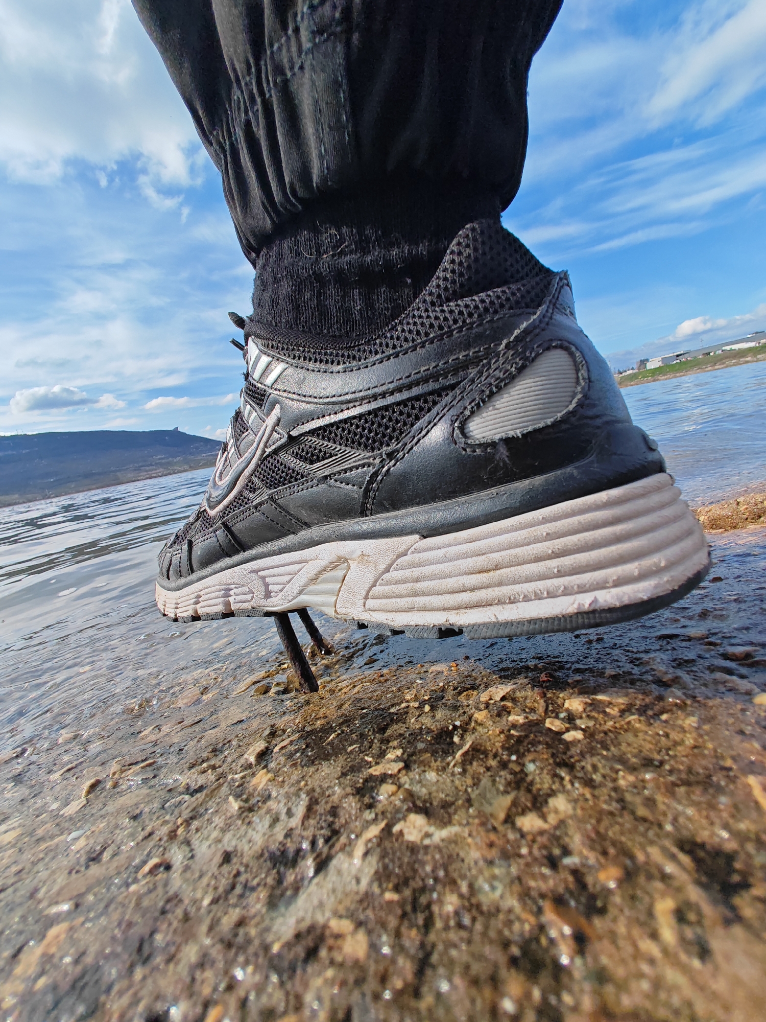 A low-angle close-up shot captures a black athletic shoe with a thick, ridged white sole resting on a textured rock surface partially submerged in water. The shoe is attached to a leg clad in black pants and a black sock. A rusty metal rod protrudes from the rock near the shoe. The body of water extends into the background, with a distant shoreline and a mountain range visible under a blue sky with scattered clouds. Based on the landscape and architecture, the location is likely in or around the city of Shumen, Bulgaria, given the presence of buildings on the skyline and the distinct mountainous terrain of the region.