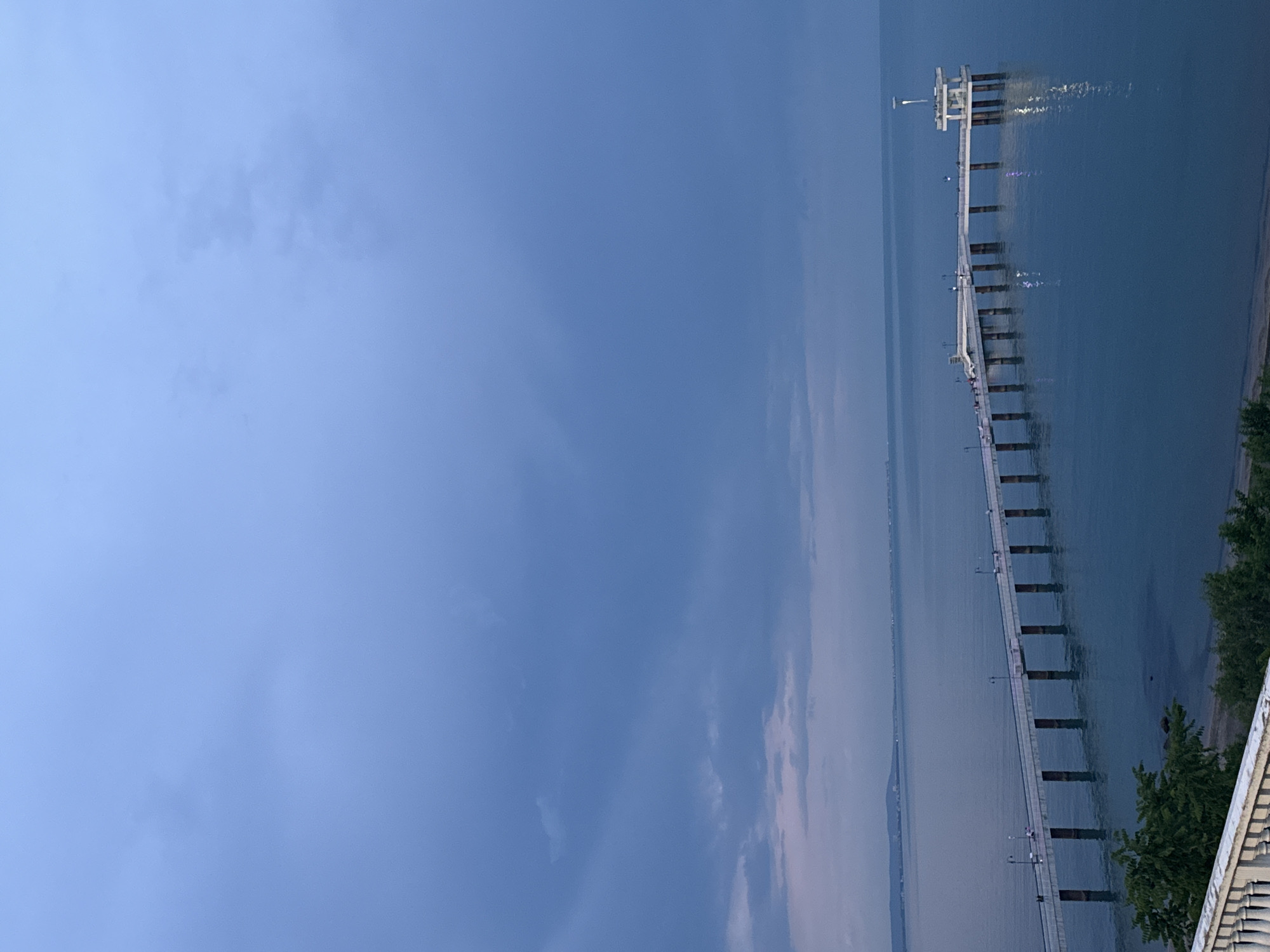 The image shows a pier extending into the sea in Burgas, Bulgaria. The sky is cloudy. The pier is constructed of concrete. There are some people visible on the pier. Trees and a building can be seen on the shoreline next to the pier.