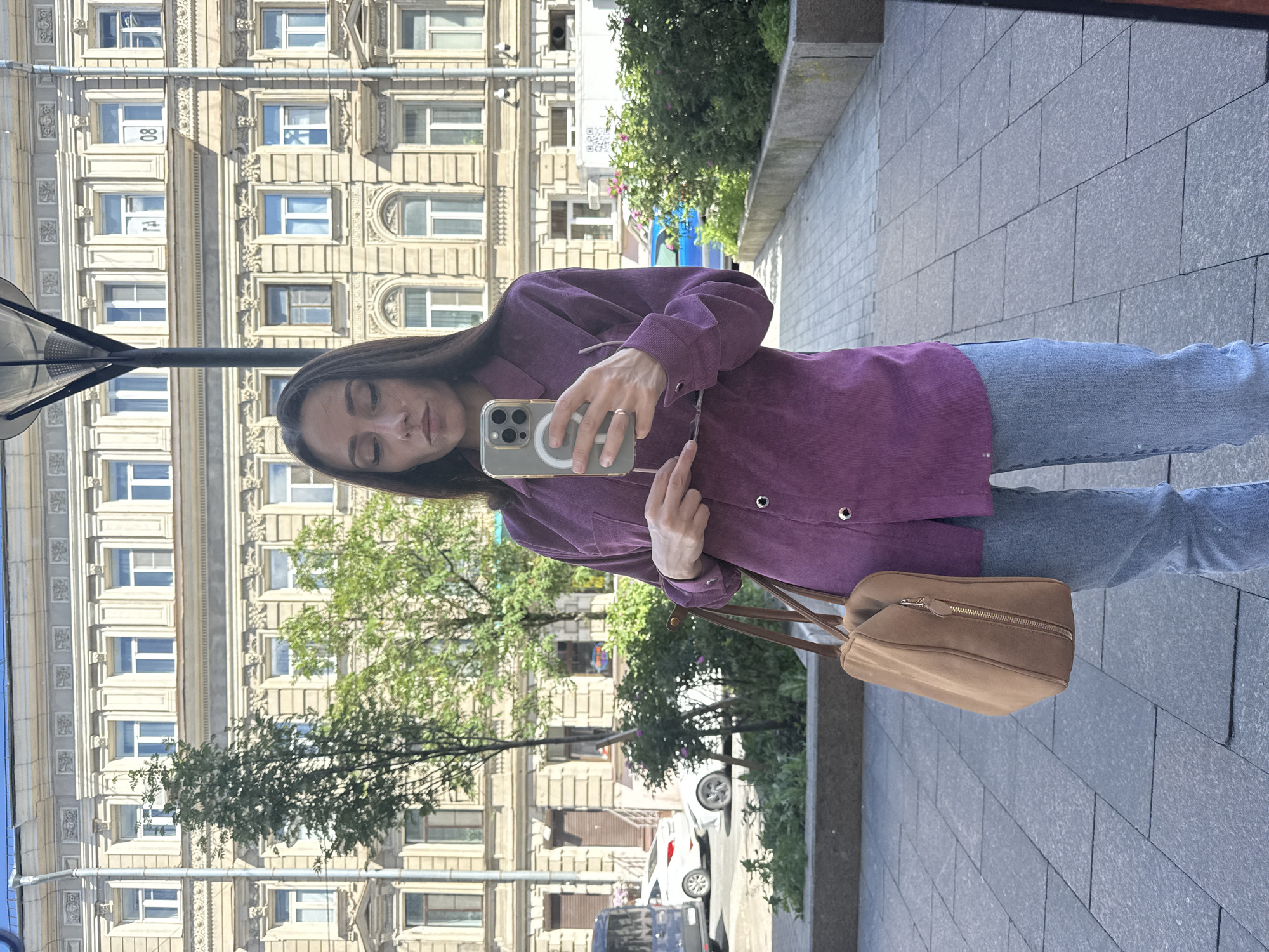 A woman with long dark hair is taking a mirror selfie in front of a building in Saint Petersburg, Russia. She is wearing a purple corduroy button-up shirt and light blue jeans. She is holding a smartphone in her right hand and a pair of sunglasses in her left hand. A brown suede shoulder bag is slung over her left shoulder. The building behind her is a multi-story structure with ornate architectural details and many windows. The sidewalk in front of the building is paved with gray tiles. Trees and some greenery are visible in the background, as well as cars parked on the street. The overall scene suggests an urban setting on a sunny day.