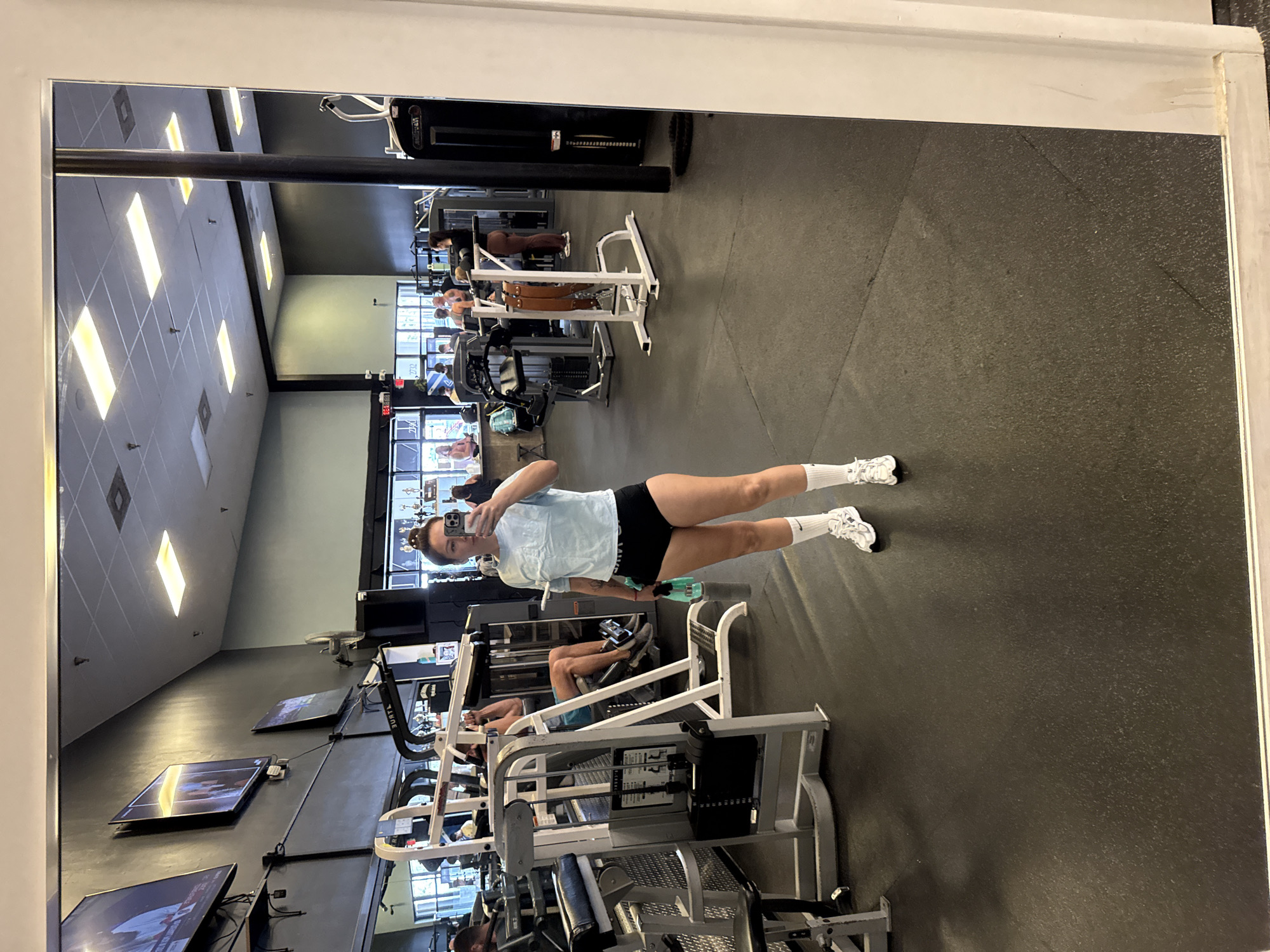 The image is a reflection in a mirror at a gym. A woman is standing in the foreground, taking a selfie with her phone. She is wearing a light blue cropped t-shirt, black athletic shorts, white socks, and white athletic shoes. She is holding a green water bottle in her left hand. In the background, several other people are visible using gym equipment, including weight machines and treadmills. The gym appears to be well-lit with fluorescent ceiling lights and has a dark rubberized floor. There are also several televisions mounted on the wall. The overall environment suggests a typical fitness facility. The image does not provide any information about the specific location in Key West, United States of America.