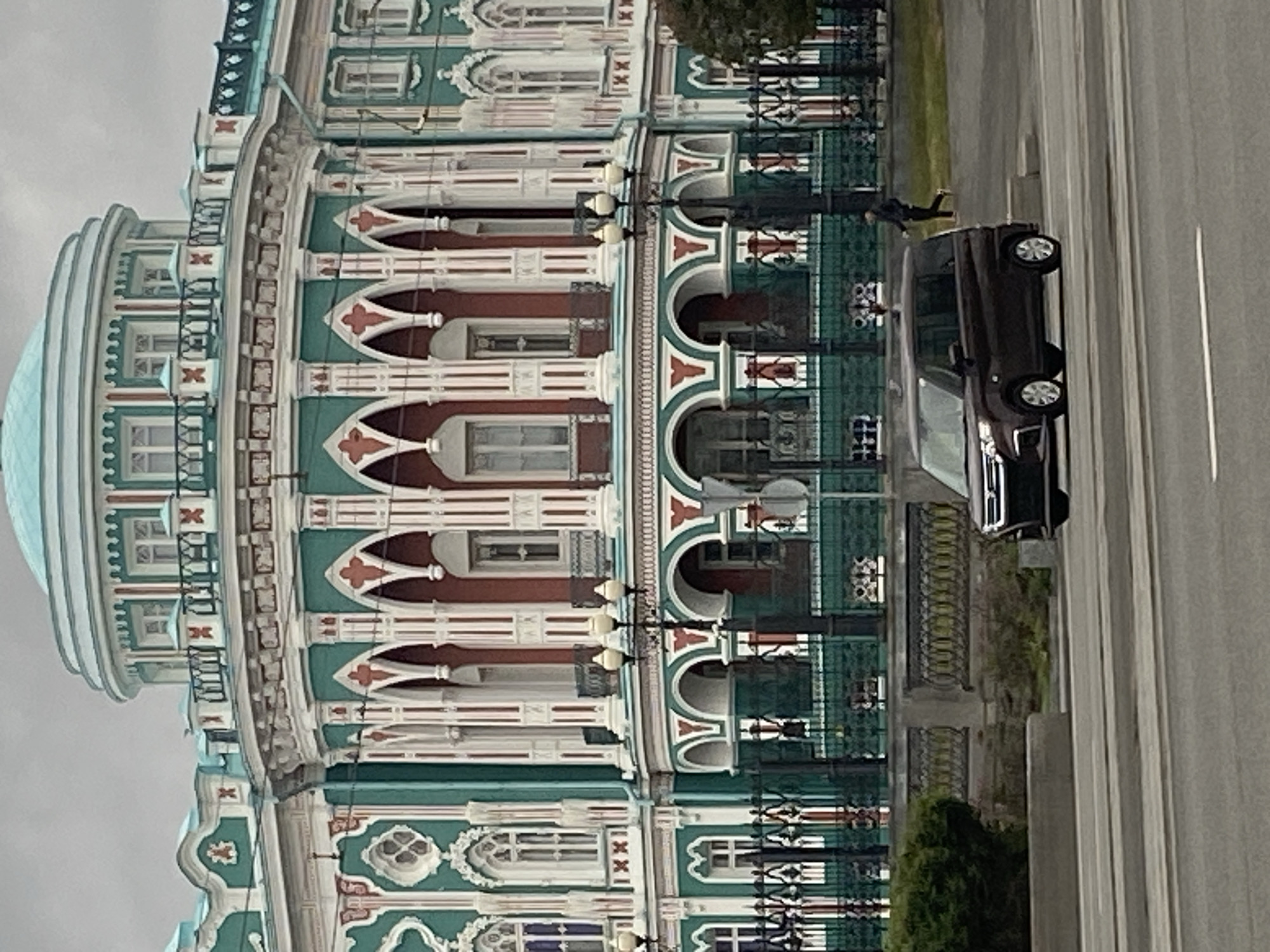 The image captures the facade of the Sevastyanov House, a landmark building in Yekaterinburg, Russia, known for its elaborate architectural style with a prominent dome and intricate ornamentation. The building displays a mix of green, white, and red colors. Next to the building, a dark-colored car is parked on the side of the road. A pedestrian is captured in motion, seemingly running or jumping near the car. The surrounding environment includes a road and some greenery, like bushes, suggesting an urban setting. The sky is overcast.