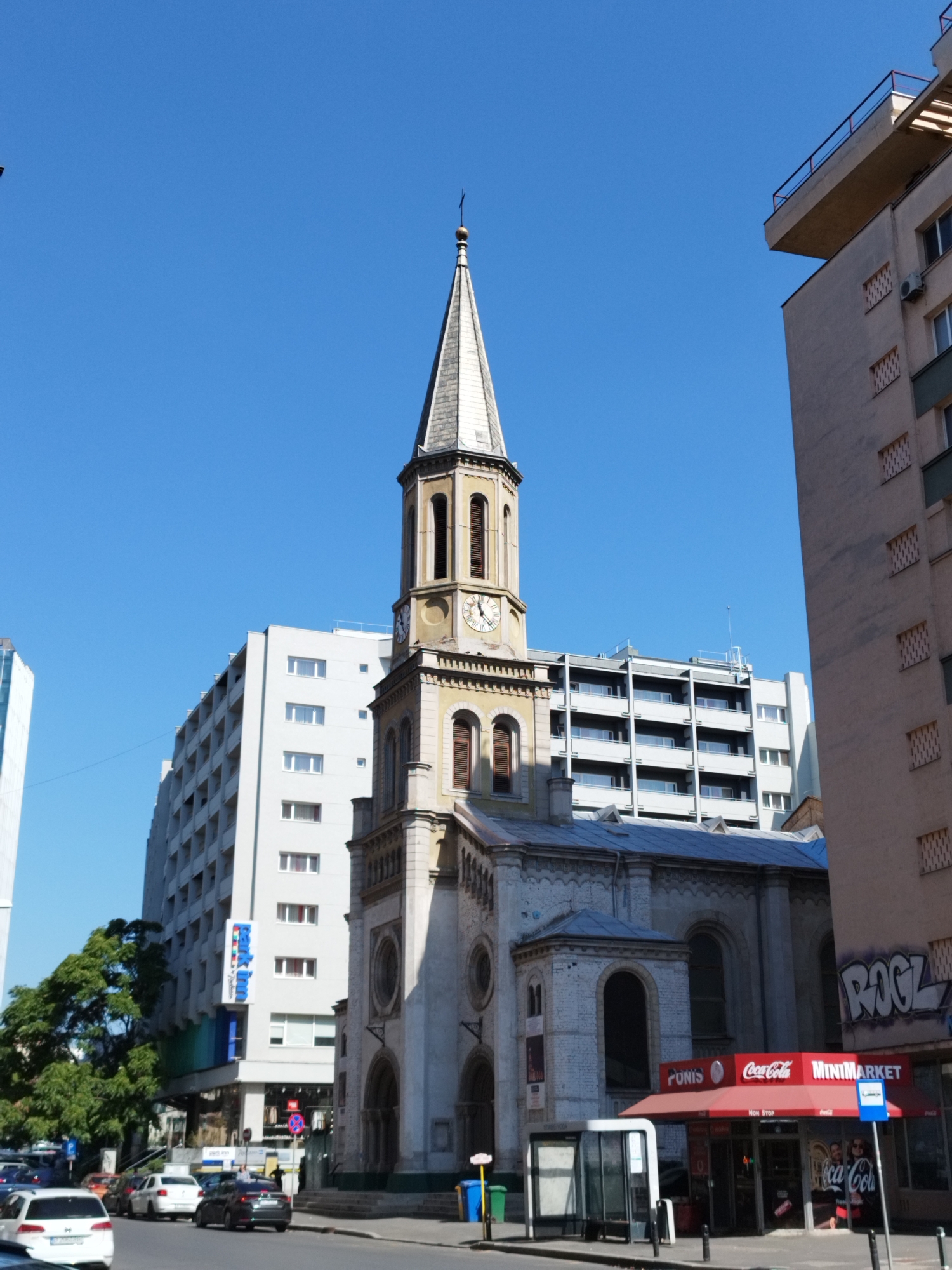 The image depicts a street scene in Bucharest, Romania, featuring a church with a prominent steeple and several multi-story buildings. The church, with its white brick facade and arched windows, stands to the right of a taller, light-colored apartment building. A clock is visible on the church tower. To the left of the church is another, more modern building with a repeating pattern of balconies. In the foreground, cars are parked along the street, and a bus stop shelter is present. A minimarket with a red awning displaying "Coca-Cola" and "Minimarket" signs is situated at the base of the church. Graffiti is visible on the side of one of the buildings. The sky is clear and blue, indicating a sunny day. The overall scene suggests an urban environment with a mix of old and new architecture.