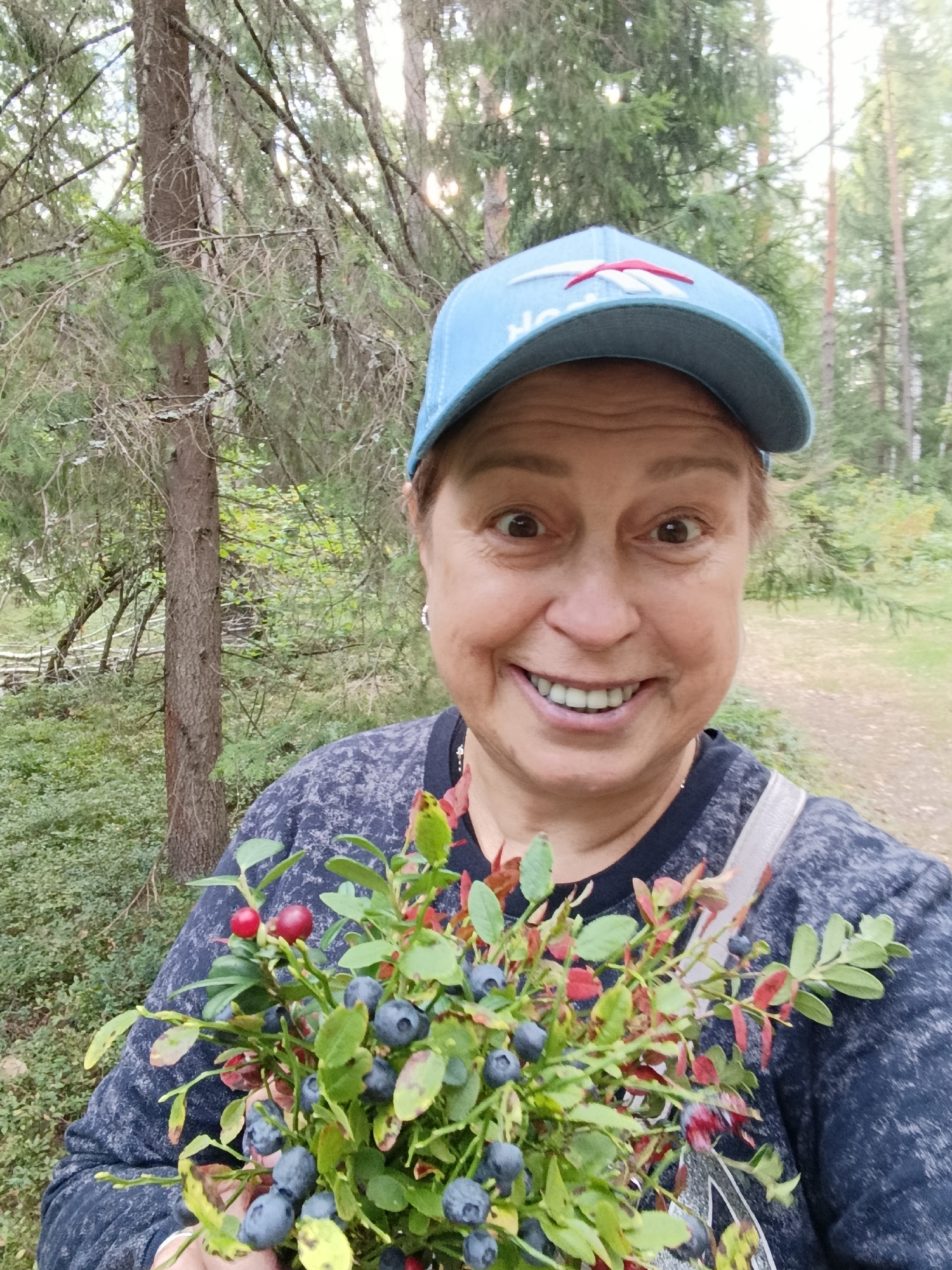A woman with fair skin and brown eyes, wearing a blue baseball cap and a dark blue sweatshirt, holds a bouquet of blueberries and two red berries. She has a broad smile, revealing her teeth. The background is a forest, with trees and green foliage. The overall scene suggests an outdoor activity like berry picking. The location is not specified in the image, but the prompt states it is in the city of Бордуки, Russia.