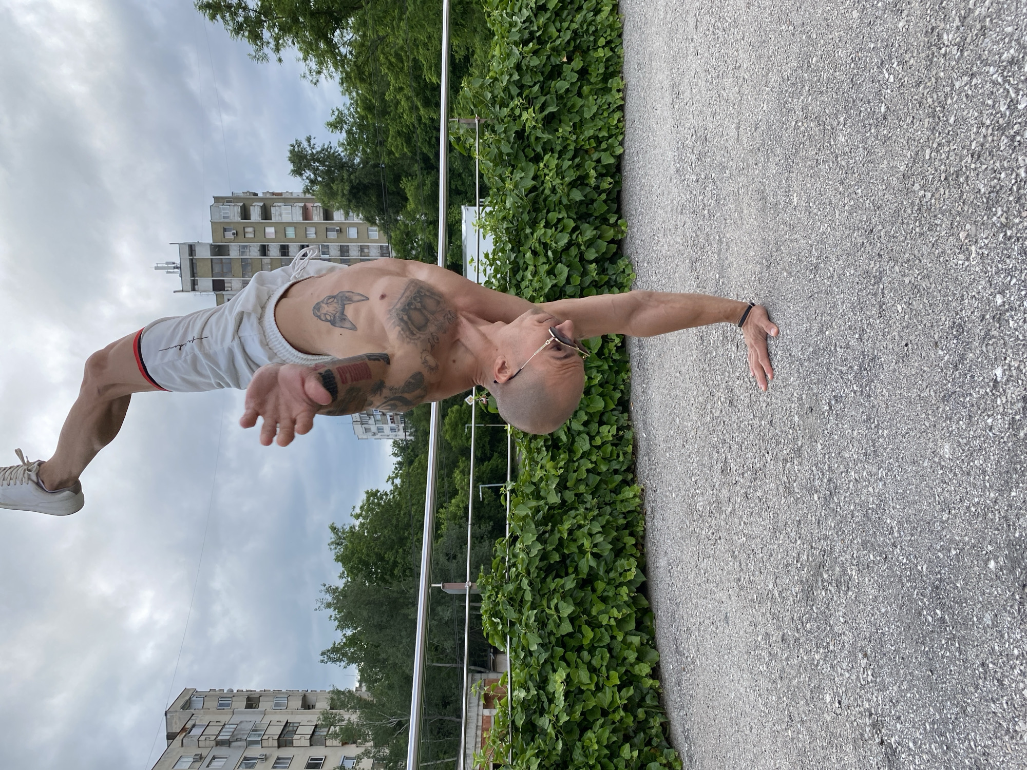 The image captures a man performing an advanced calisthenics exercise outdoors in Pleven, Bulgaria, under an overcast sky. The man, who is bald and wearing sunglasses, is shirtless, showcasing a muscular physique and extensive tattoos across his chest and arms, including a stylized head on his left pectoral. He is dressed in white athletic shorts with red and black side stripes and white sneakers.
He is captured mid-movement, holding his body horizontally, parallel to the ground, in what appears to be a demanding one-arm plank or front lever variation. His body is taut, demonstrating exceptional strength and balance, with his right hand seemingly planted on the ground for support.
The setting is an urban or suburban outdoor area. To the man's side and extending into the middle ground, there's a dense wall of green foliage or bushes, possibly ivy, growing along a silver metal railing. The ground surface where he places his hand is a light grey, coarse-grained material, resembling asphalt or concrete pavement. In the background, behind the railing and greenery, several tall, older residential buildings with numerous windows and balconies are visible, interspersed with green trees. The sky is predominantly cloudy, suggesting a diffused, soft light rather than direct sunlight. The scene depicts a moment of intense physical discipline within a public environment. No discernible text is visible.