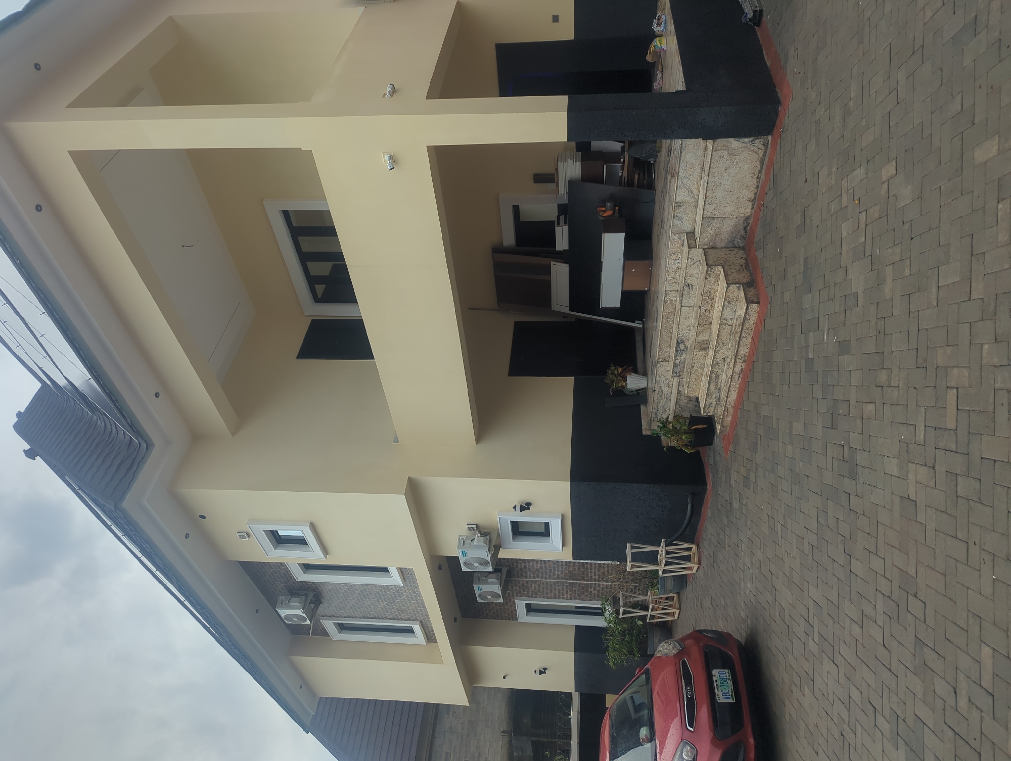 A two-story modern house in Gwarinpa, Nigeria, features a cream-colored facade with black accents on the lower level. The house has multiple windows and balconies, with an overhang above the entrance. Two air conditioning units are visible on the exterior wall. A red Kia car is parked on the patterned brick driveway in front of the house. Several wooden decorative stands are placed near the entrance, and some construction materials are stacked on the porch area. The sky is overcast.