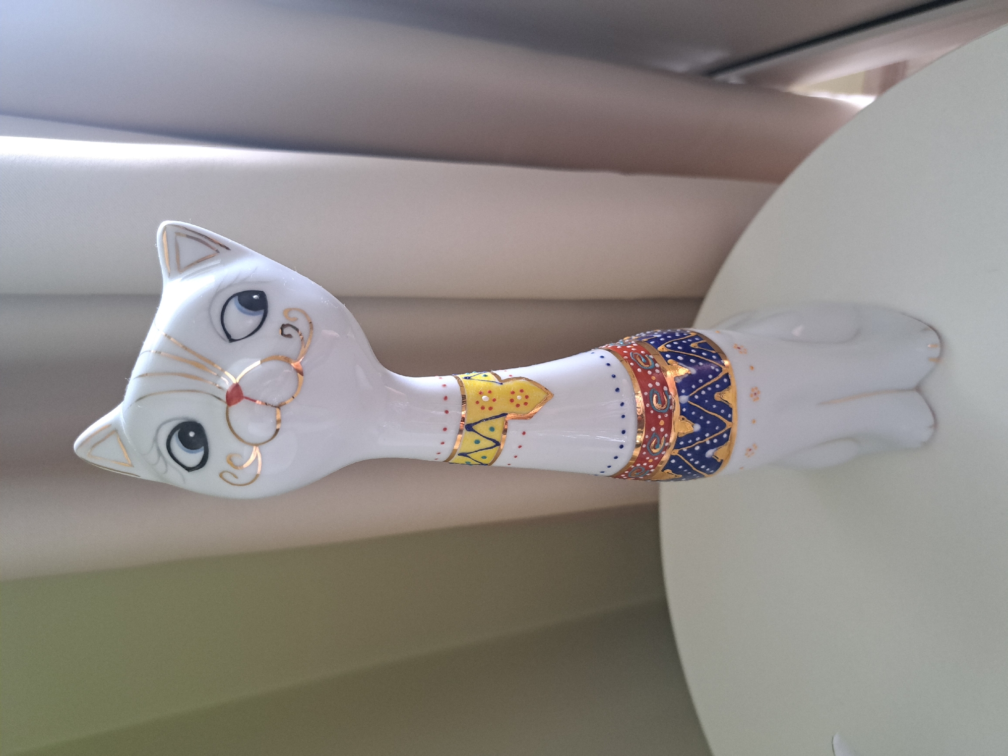 A white ceramic cat figurine with gold accents is positioned on a white circular table. The figurine has a long neck, large blue and black eyes with gold eyelashes, and painted gold whiskers and mouth. It wears decorative bands around its neck and torso in yellow, red, and blue with gold trim. The background consists of beige and gray curtains. The scene does not provide any information about the city of Irkutsk, Russia, or any specific location within it. There are no people or actions depicted in the image.