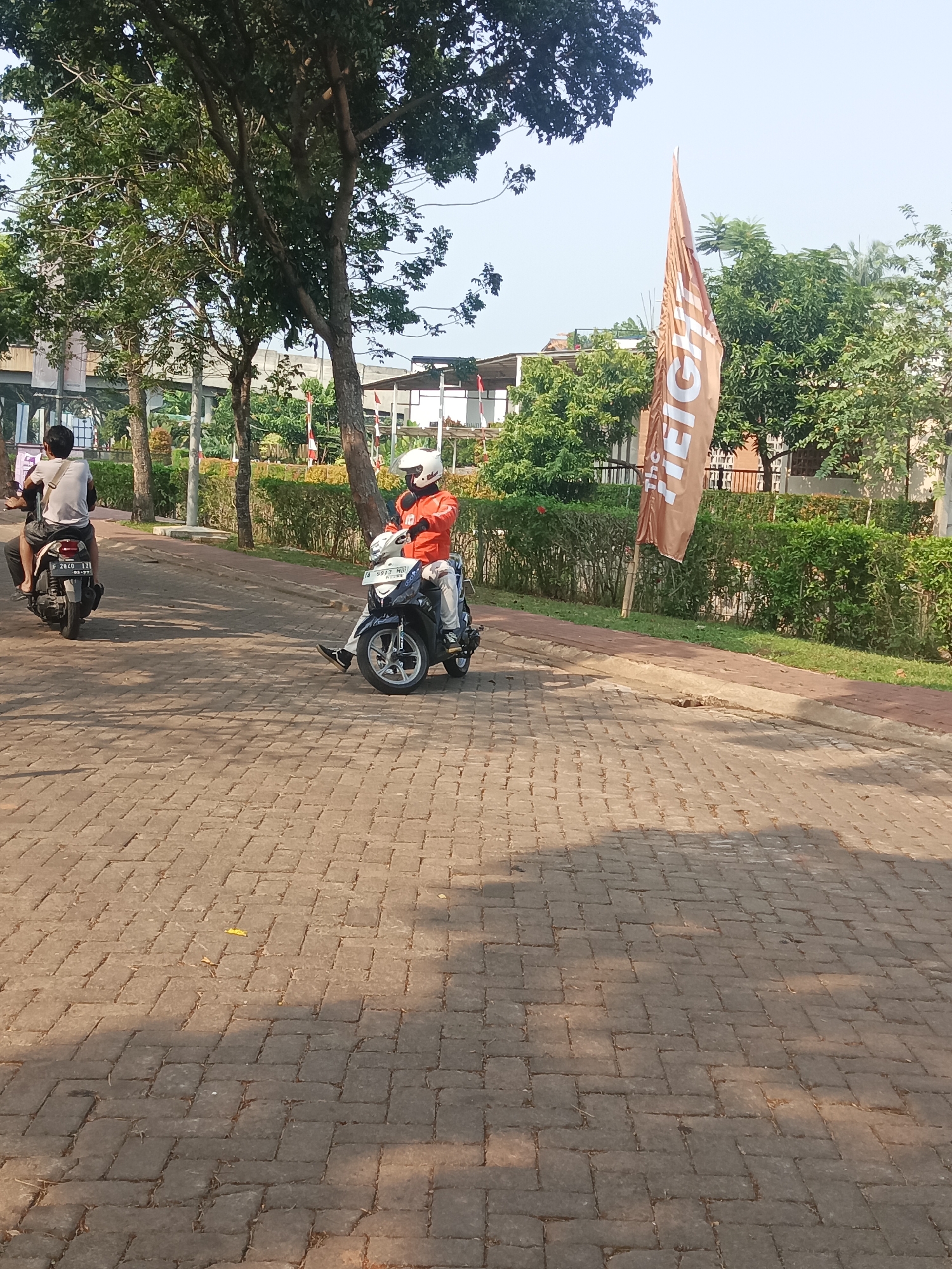 In South Tangerang, Indonesia, two individuals are observed riding motorcycles on a paved brick road. The first motorcyclist, wearing an orange jacket and a white helmet, is positioned in the foreground, looking forward and appearing to be in motion. The second motorcyclist, wearing a white t-shirt and dark pants, is visible further down the road, also appearing to be moving. The surrounding environment features trees, lush green bushes, and buildings in the background. A brown flag with white lettering, possibly advertising a business, stands prominently near the right side of the frame. The scene is illuminated by natural daylight, casting shadows on the brickwork.