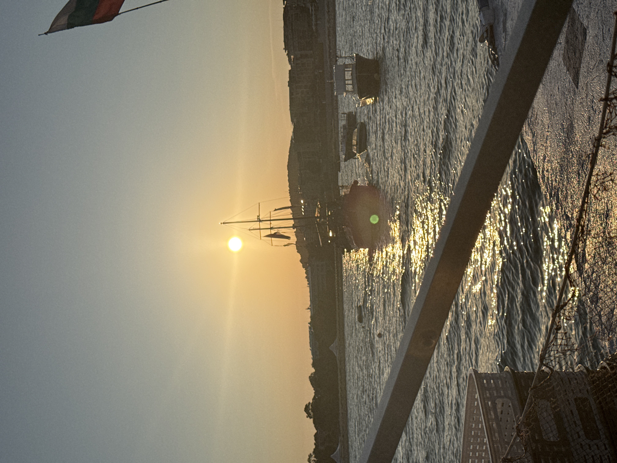 This image captures a serene sunset over the Black Sea in Primorsko, Bulgaria. The sun, a bright orb, casts a golden glow across the water, creating shimmering reflections. In the foreground, a portion of a boat's railing and netting are visible, suggesting the perspective is from a vessel or pier. Several boats are dotted across the water, including a larger sailing ship with masts and rigging, and smaller motorboats. In the distance, the coastline is lined with buildings and trees, silhouetted against the warm sky. A Bulgarian flag flutters on a pole to the right, adding a national element to the scene. The water appears calm with gentle ripples. The overall atmosphere is peaceful and picturesque.