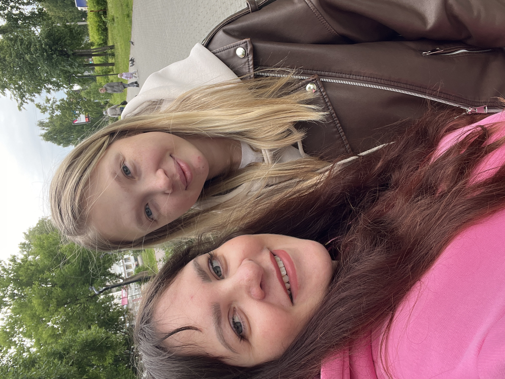 An outdoor selfie-style photograph, taken in the city of Chaikovsky, Russia, features two women posing in the foreground. The woman on the left, positioned closer to the camera, has long, dark brown hair and is wearing a bright pink hooded sweatshirt. She is smiling, revealing her upper teeth, and has light-colored eyes. A small reddish mark is visible on her forehead above her left eyebrow. The woman on the right, standing slightly behind, has long, blonde hair and is wearing a light-colored hooded sweatshirt beneath a dark brown faux leather jacket with silver zippers and snap buttons. She is also smiling with light-colored eyes.
The background depicts a public park or pedestrian area with a paved path extending into the distance. Numerous green trees with dense foliage and green grass line the path, suggesting a warm season. In the mid-ground, a black lamppost is visible on the right side of the path, and a red and white sign is partially visible on the left. Faint building structures are discernible in the far distance through the trees. Several indistinct figures are visible walking on the path further back, including adults and a small child. Multiple pigeons are present on the paved surface. The sky is overcast, indicating diffused daylight.