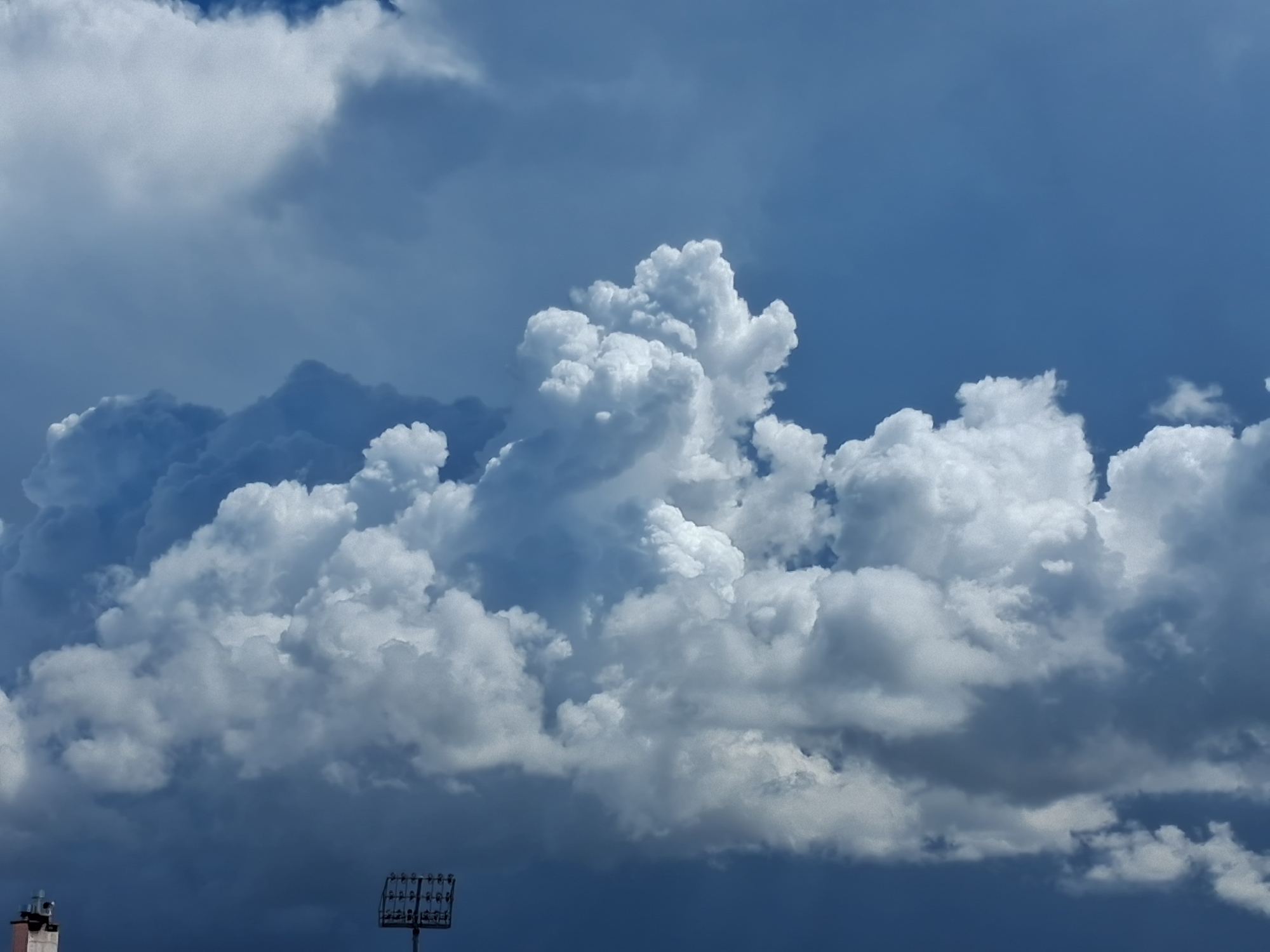 The image captures a sky filled with puffy, cumulus clouds, varying in size and density. The clouds transition from bright white to dark grey, suggesting different levels of sunlight penetration and shadow. The sky ranges from a clear, lighter blue to a deeper, darker blue. In the lower part of the image, on the left is the top of a building, possibly a chimney. On the right, there is what appears to be a utility pole or a structure with rectangular lights or components. Due to the lighting and the types of clouds, there may be the location of the city of Figueira da Foz, Portugal, during the day.