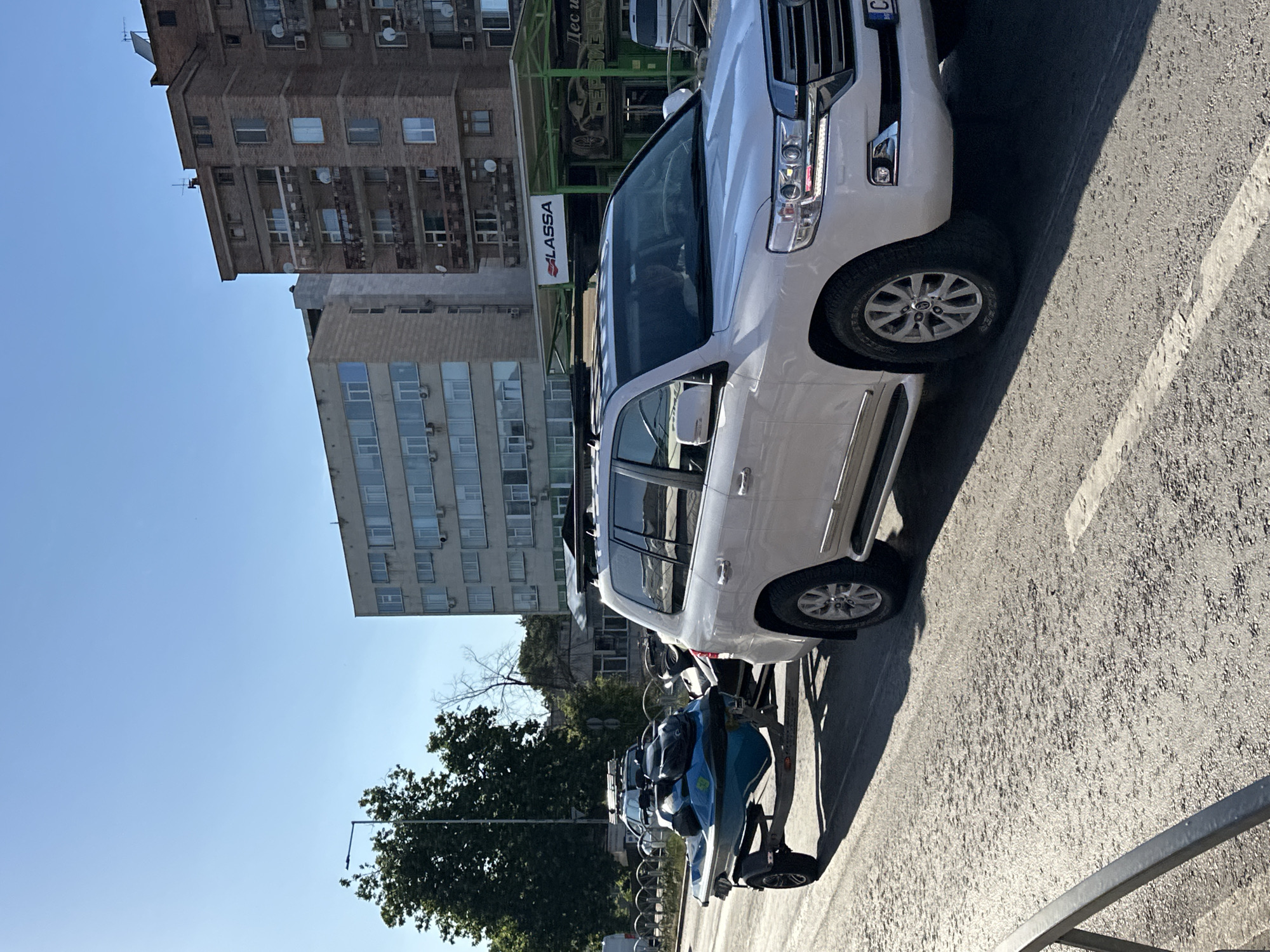 A white SUV, possibly a Toyota Land Cruiser, is parked on an asphalt road, casting a shadow. Behind the SUV, on a trailer, are several blue and black personal watercrafts. In the background, there are multi-story buildings, one appearing to be a brown brick structure, and another a lighter colored building with many windows. A green canopy structure is visible in the mid-ground. Trees with green foliage are also present. The sky is clear and blue, indicating a sunny day. The image is taken outdoors, likely in a city setting. The time of day appears to be daytime, given the clear sky and shadows. There is text visible on a red and white sign attached to the green canopy that reads "ALASSA". Another sign on the green canopy has Cyrillic text. The overall scene suggests a commercial area or a parking lot near a business.