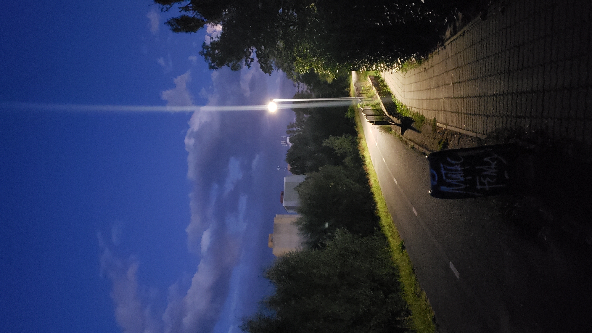 A nighttime scene in Prešov, Slovakia depicts a paved path and an adjacent bicycle lane under a deep blue sky with scattered clouds and a bright light pole. The path, made of interlocking bricks, is illuminated by the pole and cast in shadow. A bicycle lane runs alongside it, with white markings visible. To the left of the path, trees and grass line the lane, with distant buildings appearing in the background. A dark trash can with graffiti is positioned at the edge of the bicycle lane. The overall impression is one of an urban path at twilight or early night.