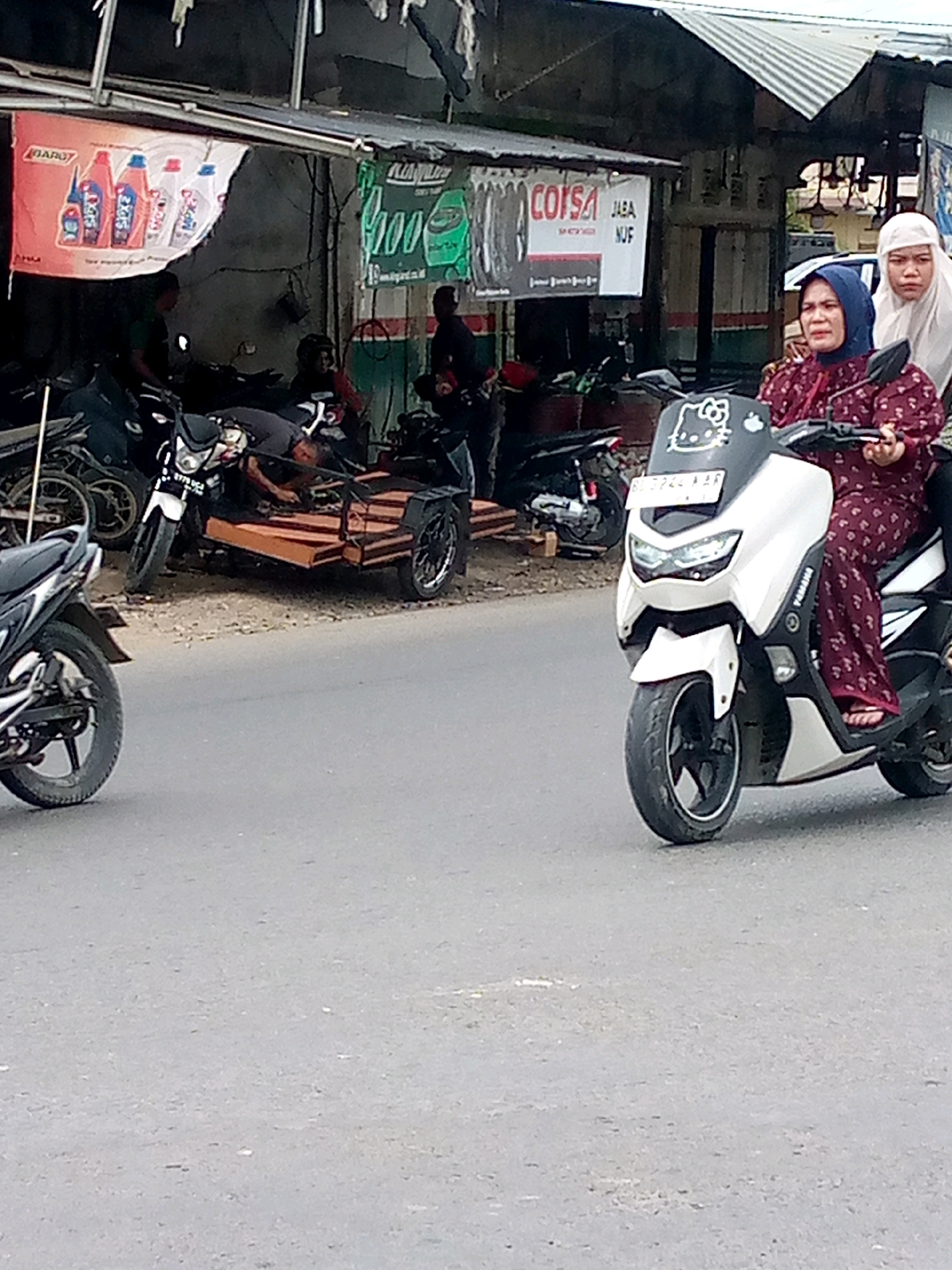 In the city of Unknown, Unknown, a side-view medium shot captures two women riding on a white scooter on a paved road. The woman in the front is wearing a maroon hijab and a matching patterned dress. She is operating the scooter. The woman behind her is wearing a white hijab. The scooter has a "Hello Kitty" sticker on the front. In the background, there are several motorcycles parked, and a shop with various banners and signs, including one with a red background and images of bottles, and another with green and white text. A trailer is attached to one of the motorcycles. There are also people visible near the shop. The environment appears to be a street scene with buildings and overhead structures.