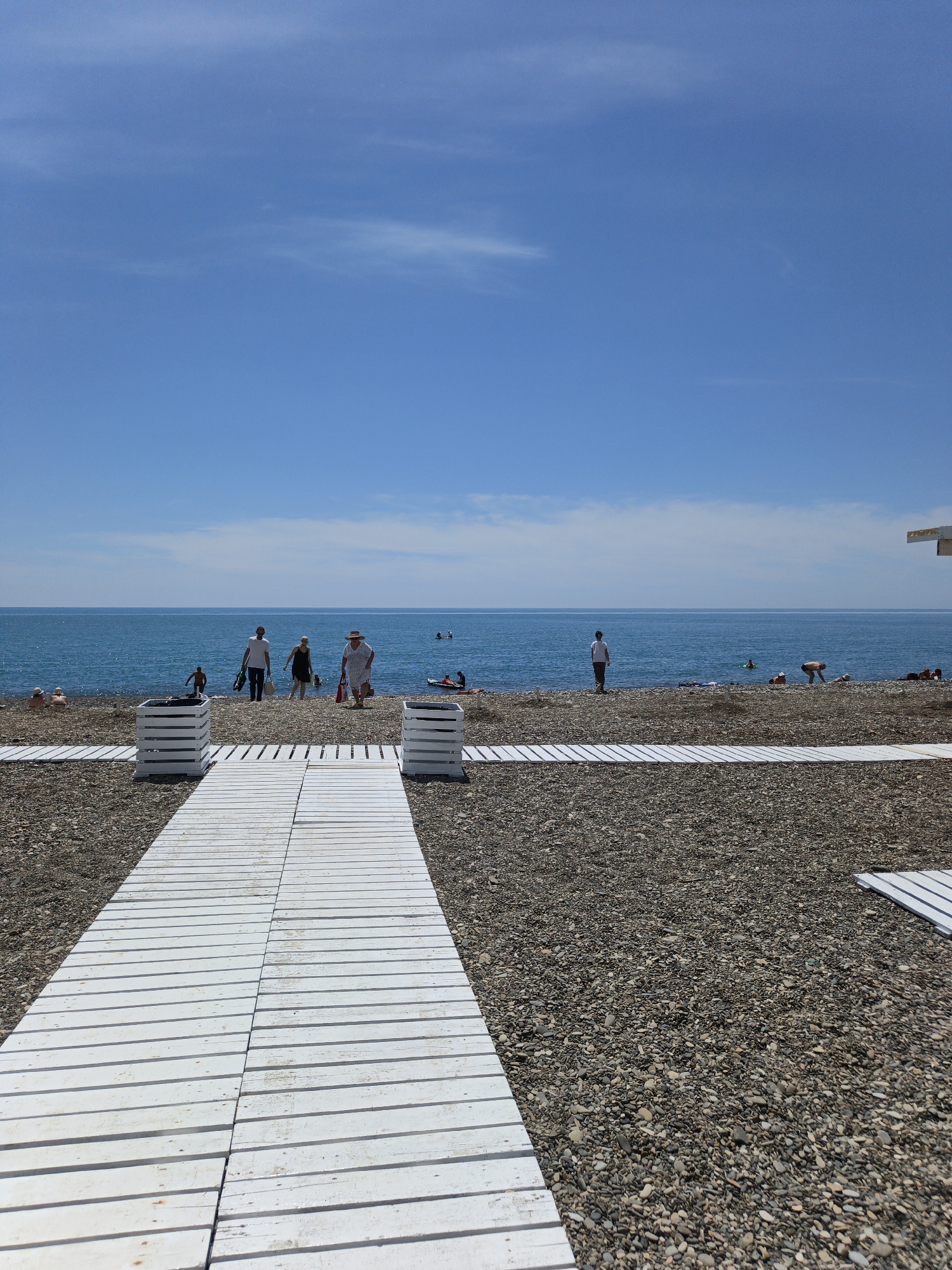 The image depicts a sunny day at a pebble beach in Krinitsa, Russia. The vast, clear blue sky occupies the upper two-thirds, with minimal wispy white clouds. A calm, deep blue-green sea extends across the middle ground to a flat horizon line. The foreground and midground consist of a coarse pebble beach, dark gray and brown in color. A central white wooden walkway, constructed from parallel slats, leads from the bottom center towards the sea. Two white wooden planter-like structures flank the walkway in the immediate foreground. Another section of white wooden path is visible entering from the bottom right.
Several individuals are present on the beach and in the water. A group of four adults is walking towards the sea along the central walkway's end, with one male carrying a black bag, a female in a dark dress, an older female in a white dress and straw hat, and a person in dark swim trunks. To the left, two individuals are seated on the pebbles near the water's edge. Further right, a person in a white t-shirt stands facing the sea, while others are swimming or lounging in the water and on the beach. One individual is paddling a small red inflatable craft near the shore on the right. The corner of a white structure is visible on the far right edge of the image near the horizon.