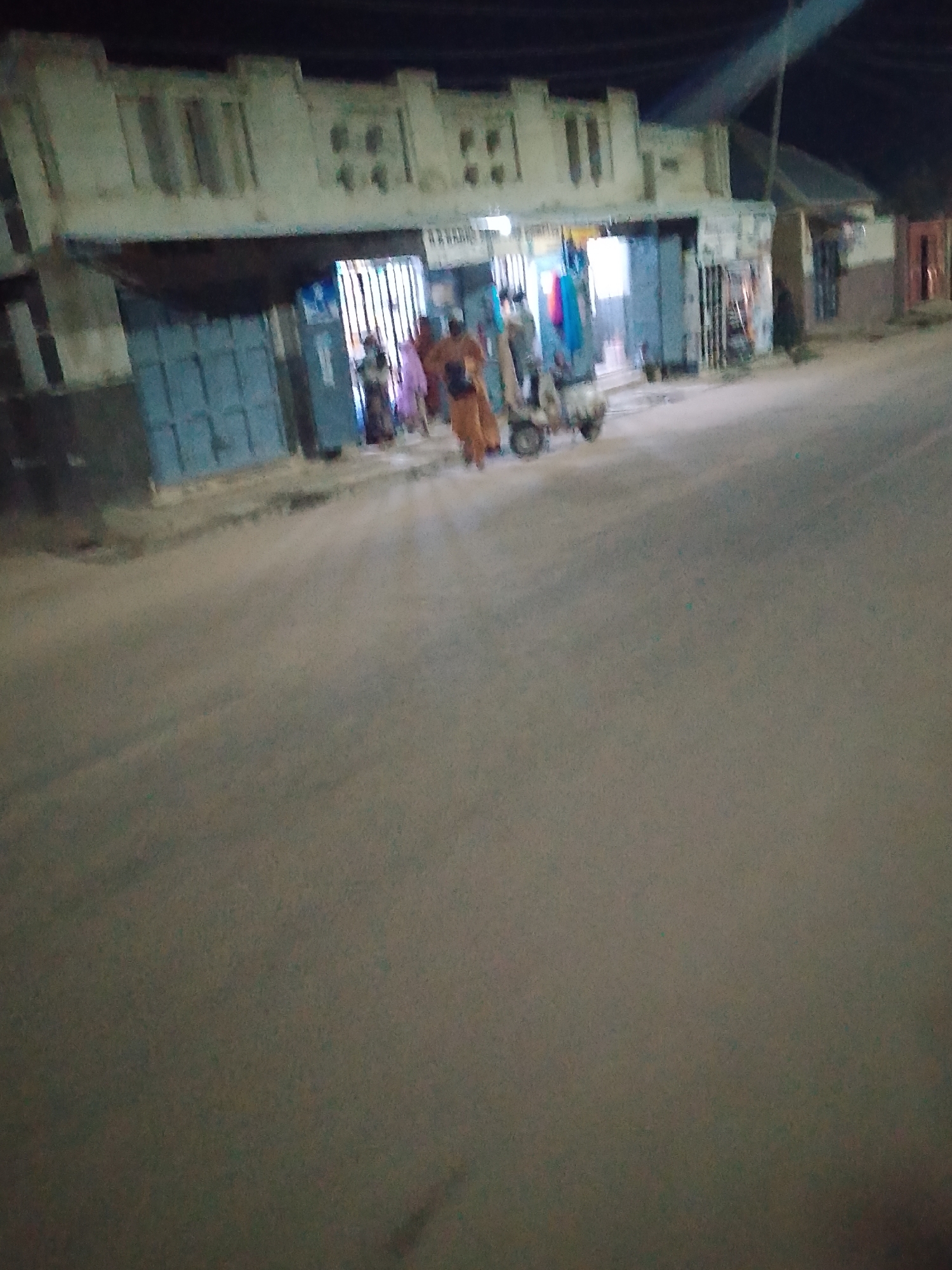 The image depicts a street scene at night in Arawa, Nigeria. Several people are present near a building with multiple open storefronts or stalls. Some individuals are dressed in traditional attire. A motorized scooter or small vehicle is visible on the street near the people. The road is paved, and street lighting casts a glow over the area. The building appears to be a commercial structure with a distinctive architectural design. The overall atmosphere suggests an evening scene in a busy area of the city.