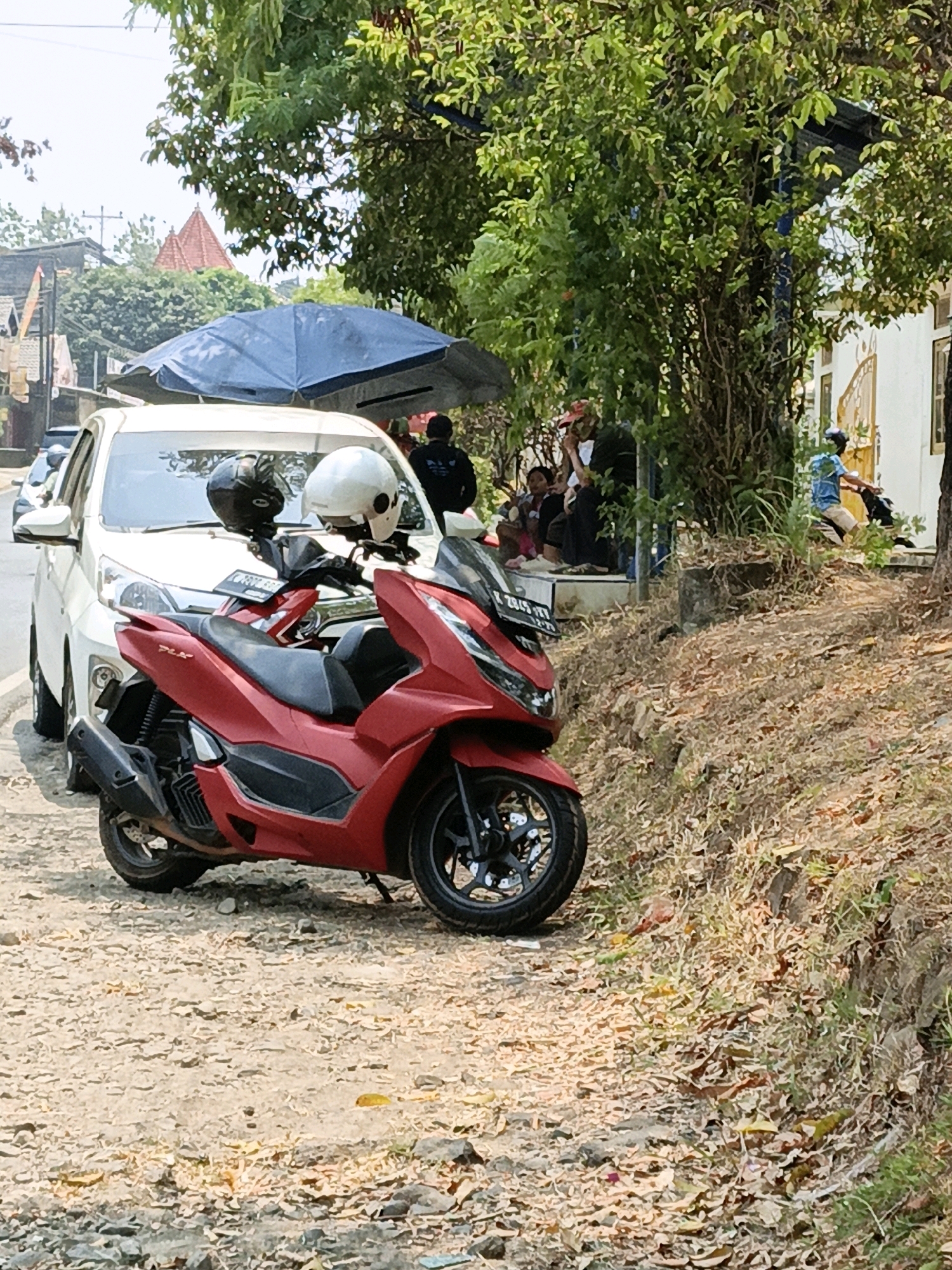 In Kuwasen, Indonesia, a red scooter with two helmets, one white and one black, rests on a dirt path in front of a white car. A blue umbrella shades the car and some people sitting on a ledge. Lush green trees and foliage line the background. A person in a blue shirt rides a motorcycle down the street. The scene appears to be a typical roadside setting on a sunny day.