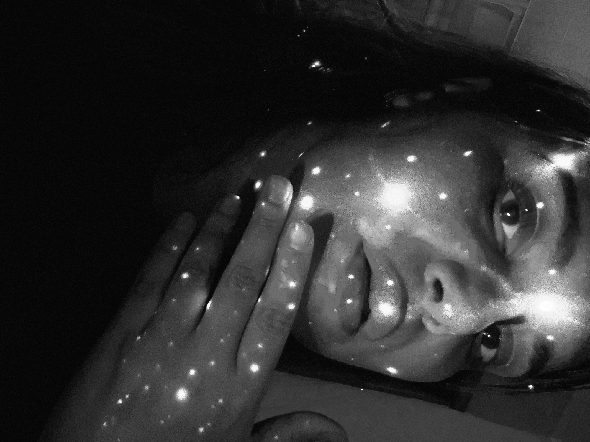 A monochromatic, close-up portrait captures a person, likely female, with their head tilted to the right, looking upwards. The primary focus is on their face and right hand, which is gently placed against the left side of their face. The scene is characterized by a stark contrast between deep shadows and numerous small, intensely bright, point-like lights. These luminous dots are scattered across the person's face—highlighting the forehead, cheekbones, nose, and lips—and continue onto their fingers and hand.
The effect of the lights resembles a starry sky, cosmic dust, or a bokeh effect, lending an ethereal and dreamlike quality to the image. The person's eyes are open and directed upwards, conveying an expression that appears calm, contemplative, or perhaps lost in thought. Their hair, visible at the top, is dark and blends into the shadowed background. The setting is dark, suggesting night or a dimly lit indoor environment. No specific location cues are visible within the image itself, though the context provided indicates the location is Chaikovsky, Russia. There is no visible text. The overall aesthetic is mysterious and artistic, emphasizing the interplay of light and shadow on human form.