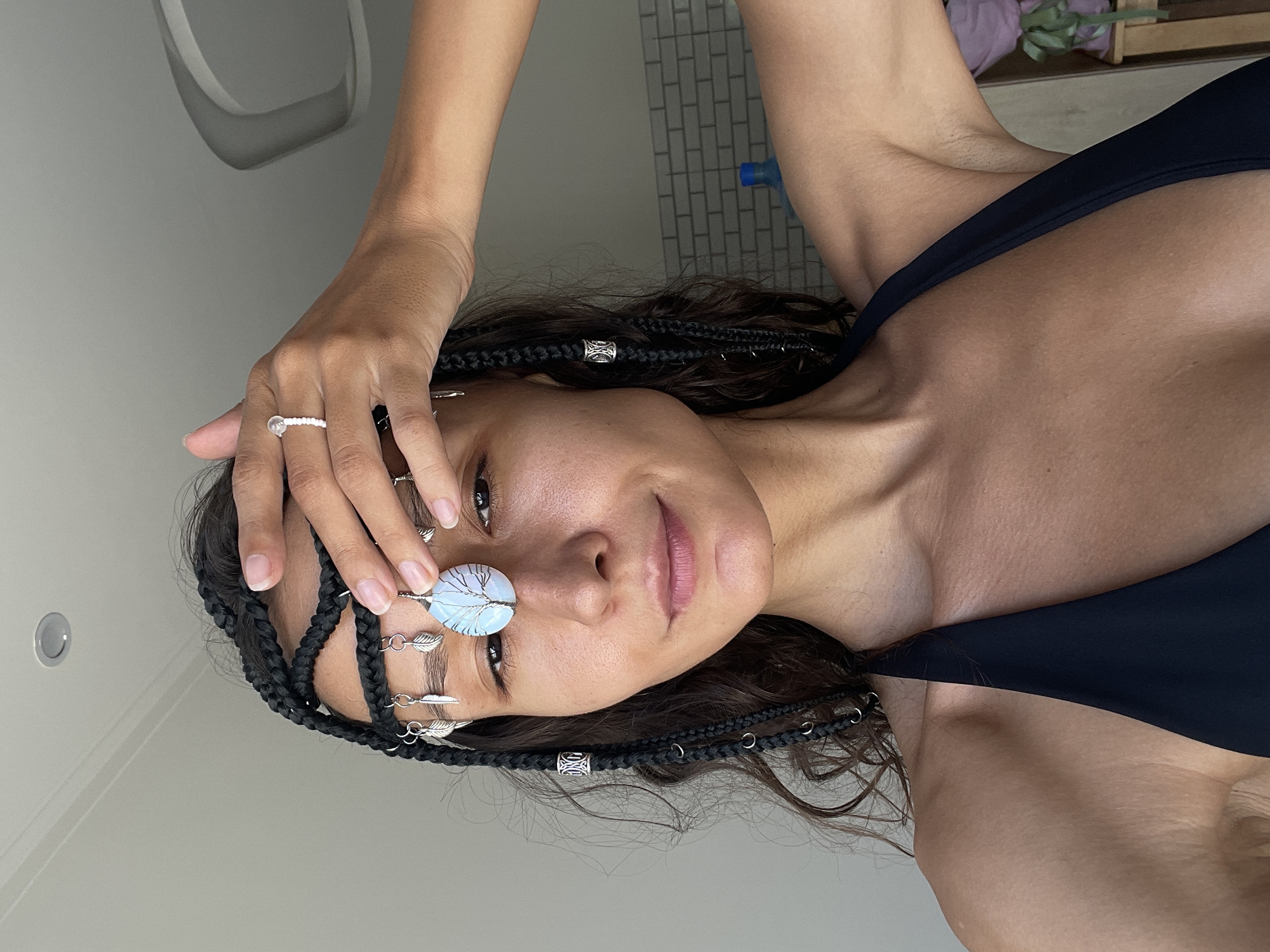 A young woman with braided dark hair and an elaborate headpiece featuring a tree-shaped pendant and leaf charms is posing for a selfie. She is wearing a black bikini top and has her right hand raised to her forehead, displaying a ring with a small white stone. The background shows a neutral-colored wall and a glimpse of a tiled surface and a blue bottle. The lighting suggests an indoor setting, possibly a home. There are no discernible elements in the image that would indicate a specific location in Mũi Né, Vietnam.