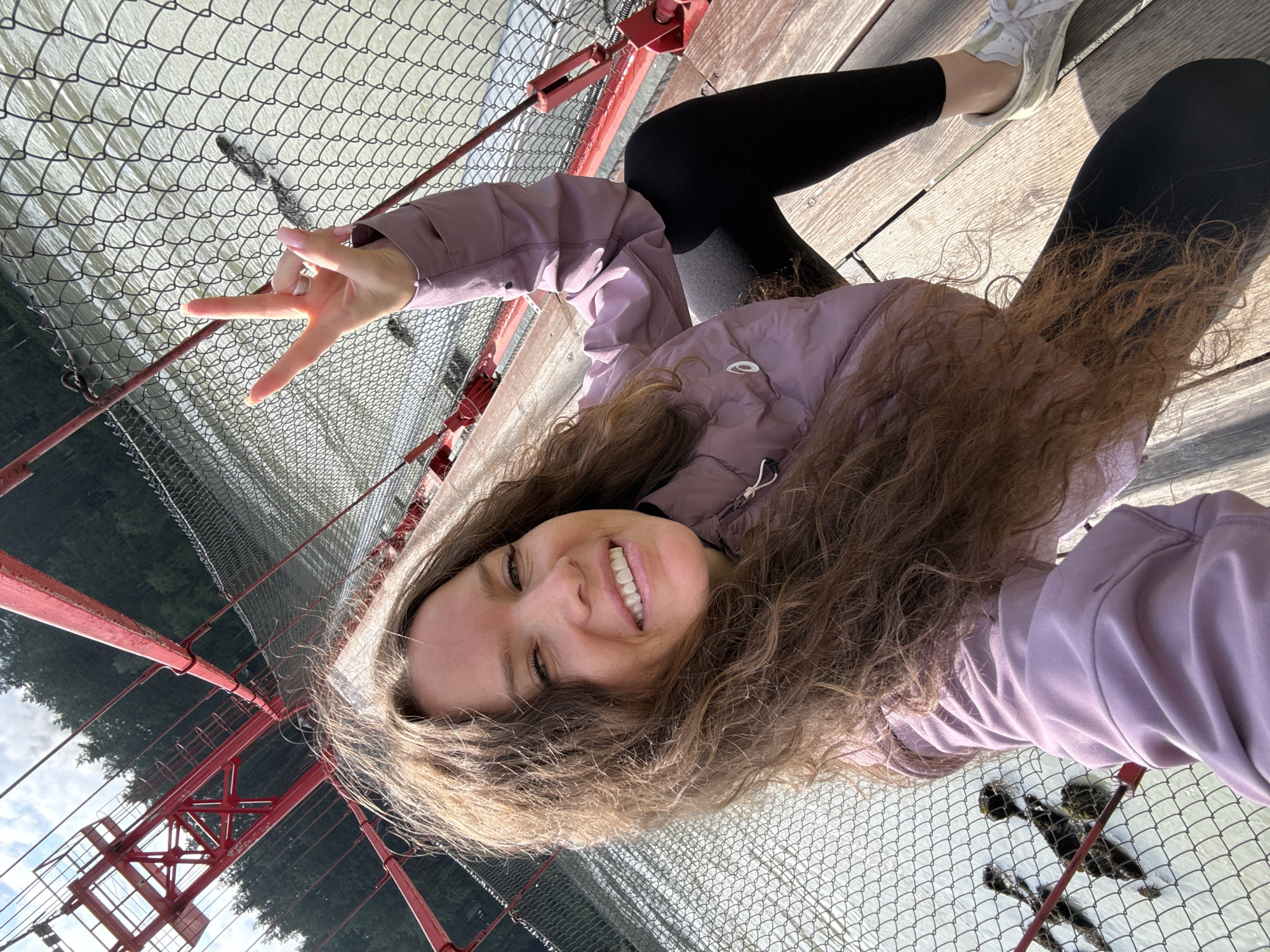 A woman with long, curly brown hair is taking a selfie on a red suspension bridge over a body of water. She is wearing a light purple jacket, black leggings, and white sneakers. Her right hand is raised, making a peace sign. The bridge has a chain-link fence along the side, and the water below appears murky. Trees and foliage are visible in the background. The image is taken from a high angle, looking down at the woman and the bridge. There is no visible indication of the location in the city of Barangol, Russia.