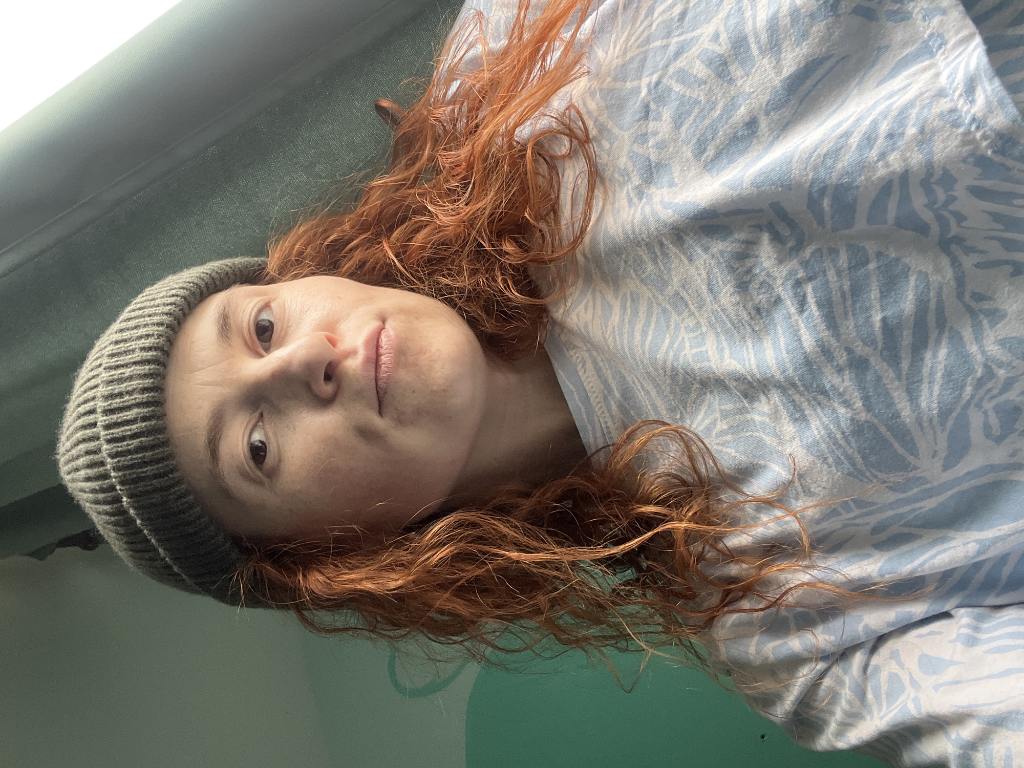 A close-up, informal portrait features a person with long, wavy, reddish-brown hair and a fair complexion, captured from an overhead perspective. The individual is wearing a grey knitted beanie and a light blue top adorned with a white pattern resembling stylized leaves or natural forms. Their head is tilted, and they are looking directly at the camera with wide, brown eyes and a neutral expression, lips slightly parted.
The setting appears to be indoors, with a solid dark teal or green surface forming the background behind the person's head and hair. This surface might be a wall or a piece of furniture. A lighter, possibly white or brightly lit, area is visible in the upper left corner of the frame. The image is oriented such that the person's head is on the left side, suggesting they are lying down or reclining.
There are no overt activities taking place beyond the act of being photographed in a casual moment. No specific time of day or weather conditions can be discerned from the image. No visible text is present. The provided location context is Chaikovsky, Russia.