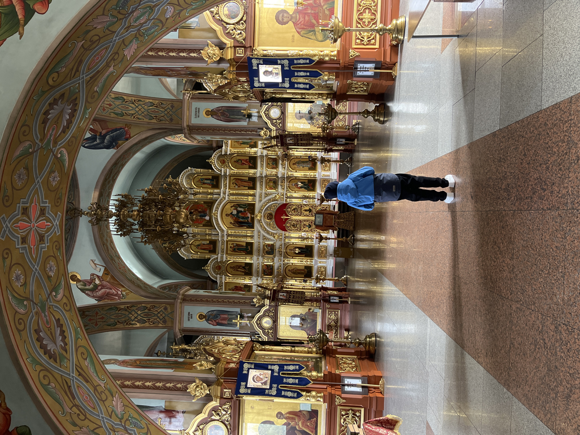 The image captures the interior of an Eastern Orthodox church, likely in Chaikovsky, Russia. The central feature is a magnificent, multi-tiered iconostasis, a wall of religious icons and paintings, richly adorned with gold gilding, intricate carvings, and decorative elements.
One person, viewed from behind, stands in the foreground, facing the iconostasis. They are wearing a blue jacket, black trousers, and white athletic shoes, adopting a posture that suggests reverence, contemplation, or prayer.
The church's architecture features high, ornate arched ceilings decorated with detailed frescoes and painted patterns in various colors, including greens, browns, and reds. Numerous icons, depicting saints and biblical figures, are displayed within the iconostasis, some featuring Cyrillic text. A blue banner with an icon and gold embroidery hangs prominently near the lower section of the iconostasis. Large brass candelabra or candle stands are visible on the floor before the iconostasis.
The floor is tiled with a contrasting design: a lighter grey and white speckled path leads towards the iconostasis, flanked by a darker, reddish-brown speckled tile in the foreground and sides. Parts of an elaborate gold chandelier are visible hanging from the ceiling. The overall scene conveys a sense of grandeur and spiritual devotion within a traditionally decorated Orthodox place of worship.