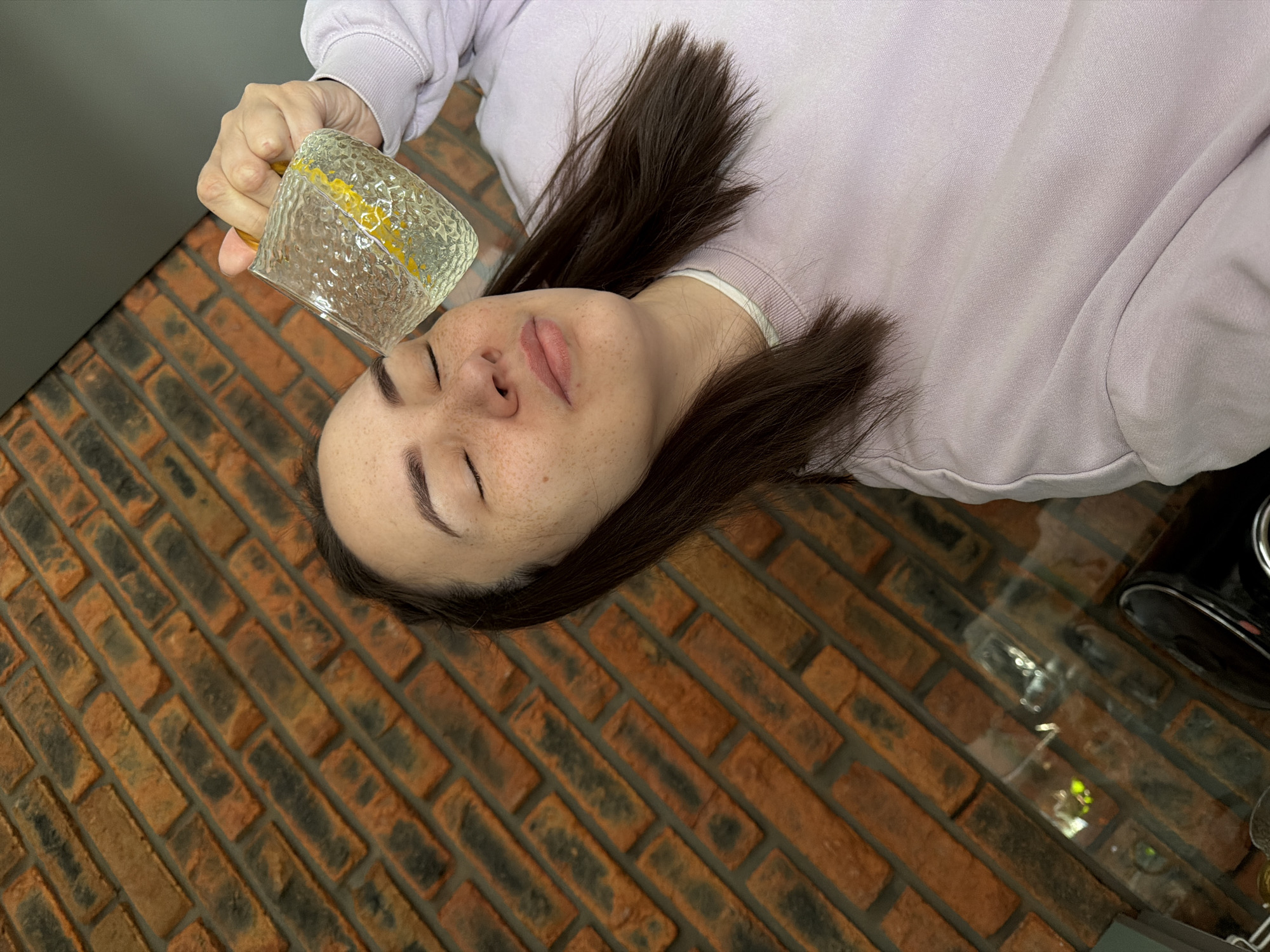 An indoor, high-angle shot captures a young woman lying on her back in what appears to be a home setting in Chaikovsky, Russia. She has dark hair fanned out around her head and noticeable freckles across her nose and cheeks. Her eyes are closed, conveying a sense of relaxation or rest. She is dressed in a light purple long-sleeved sweatshirt.
Her right hand holds a textured, clear glass mug, possibly with a yellow-tinted handle or interior, upside down directly above her forehead. The mug appears empty.
The woman is positioned on a surface covered with reddish-brown and dark gray brick-patterned tiles, laid out diagonally, which might be a floor or a large countertop. To the left, a plain dark gray wall or cabinet surface is visible. On the far right, a dark, partially reflective object, possibly a kitchen appliance, can be seen. The overall lighting is soft and even, typical of an indoor environment. There is no visible text in the image. The scene suggests a quiet moment of personal relaxation or a home remedy.