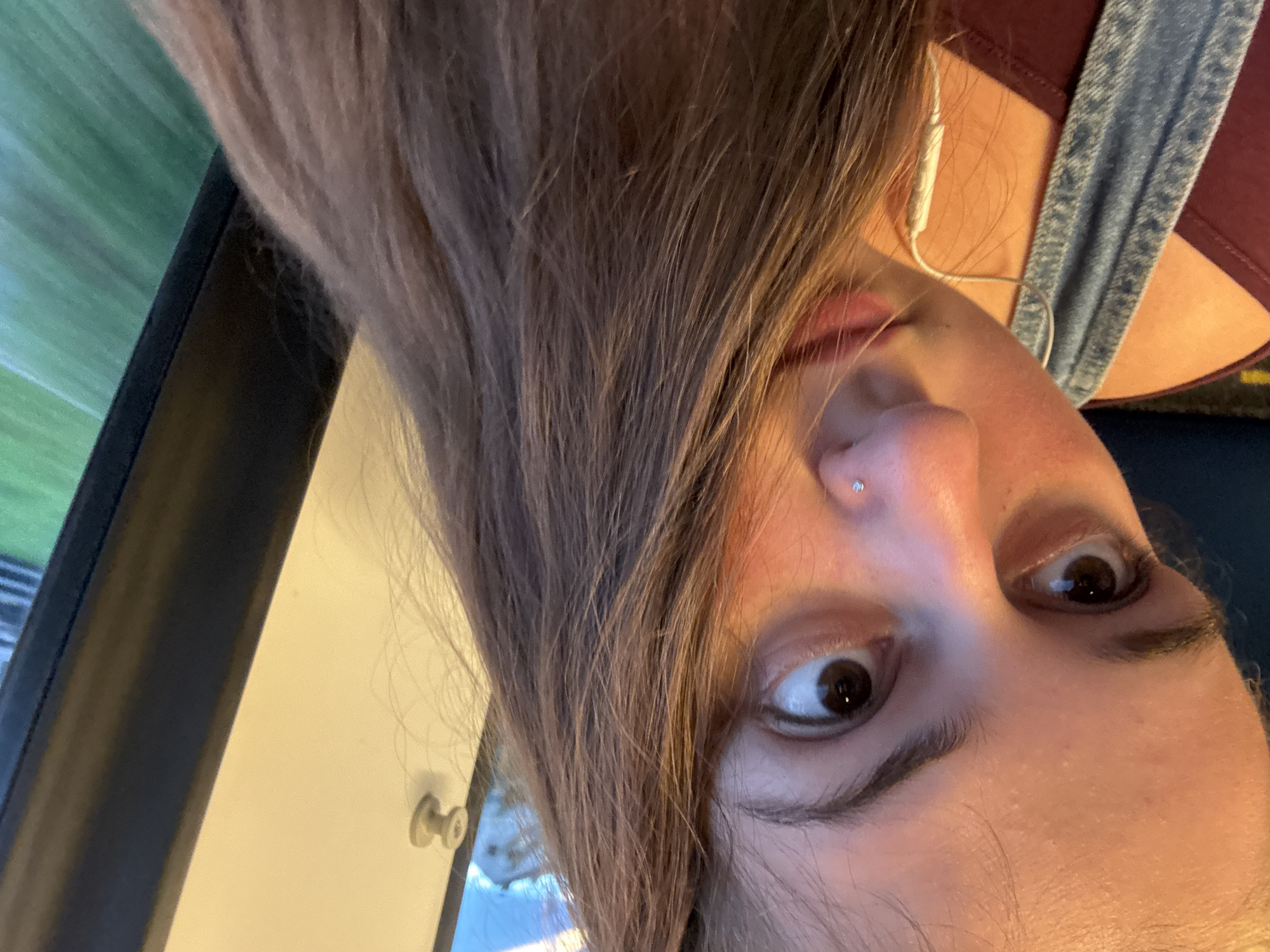 The image is a close-up, selfie-style photograph of a young woman with long, brown hair. She is wearing a dark red top and denim overalls. A white earbud cord is visible around her neck. She has a nose stud. The background shows a window with a blurry green landscape outside and a light-colored wall with a grey circular handle. The lighting suggests it is daytime. The specific location within Schopfheim, Germany, cannot be determined from the image alone.