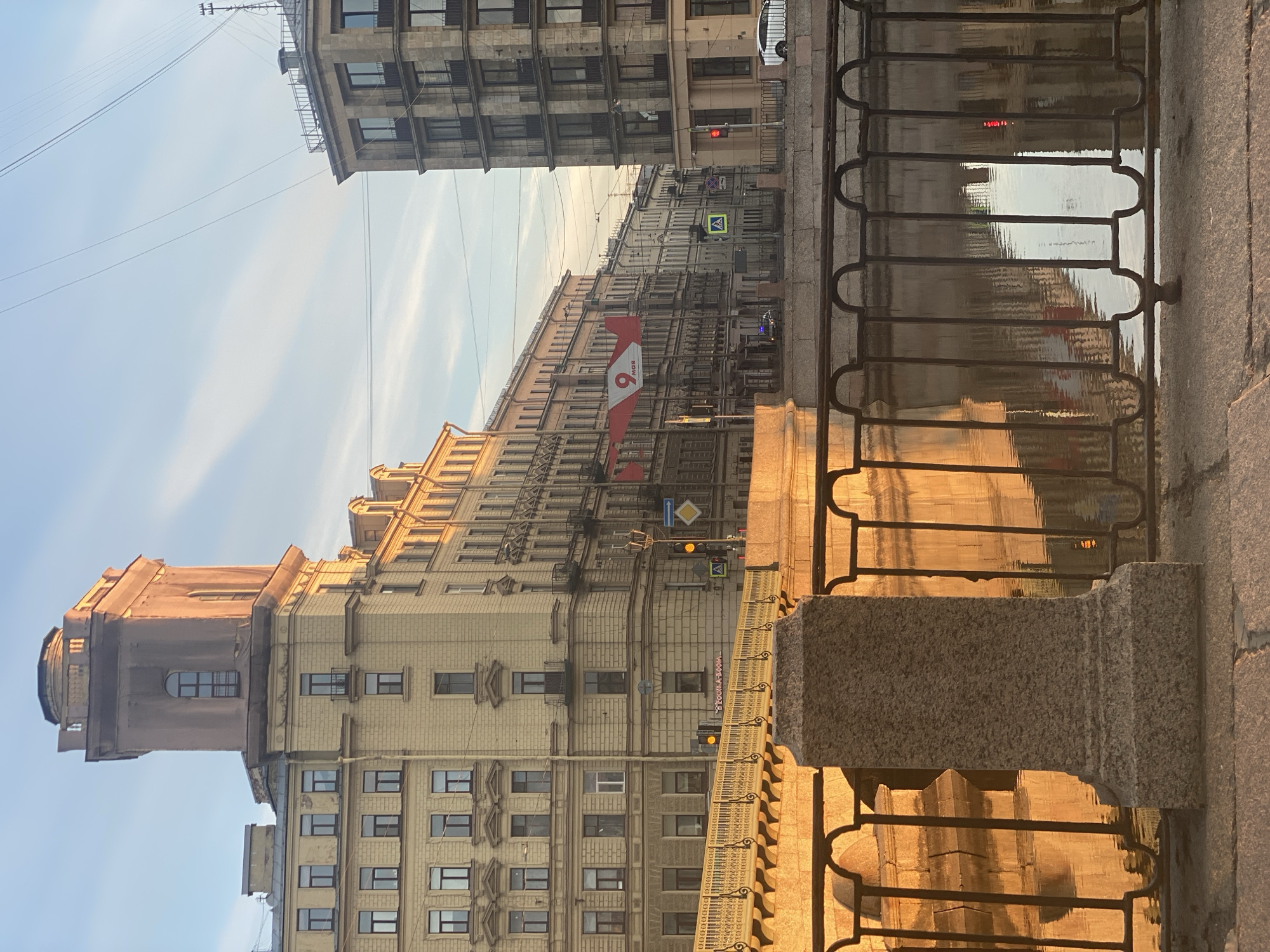 The image depicts a serene urban landscape featuring a canal flanked by historic buildings under the soft light of the golden hour. The perspective is from an embankment, with a decorative metal railing and stone balustrade in the foreground on the right.
Multi-story buildings with classical and possibly Art Nouveau architectural styles line the street on the left and in the background. The building closest on the left features a distinctive, ornate tower-like structure with a small cupola at its corner. Further along the street, a large banner is prominently displayed on another building. This banner is red and white, showing a large red number "9" with the Cyrillic word "МАЯ" (MAY) below it, strongly suggesting a commemoration of May 9th (Victory Day in Russia).
The canal, which occupies the lower right of the frame, beautifully reflects the sunlit buildings and the clear, light blue sky, creating shimmering patterns on its surface. The sky, visible above the buildings, is mostly clear with a few faint, wispy clouds.
Street furniture is visible, including traffic lights (one red, one yellow) and various road signs. Among the signs are a blue square with a white triangle depicting a pedestrian, indicating a pedestrian crossing, and a yellow diamond sign, typically signifying a priority road. A dark vehicle can be seen in the distance on the street. The warm, directional sunlight suggests either early morning or late afternoon, contributing to a peaceful and atmospheric scene. There are no people visibly engaged in activity, reinforcing the quiet ambiance. The architectural style, canals, and visible text align with the given location context of Saint Petersburg, Russia.