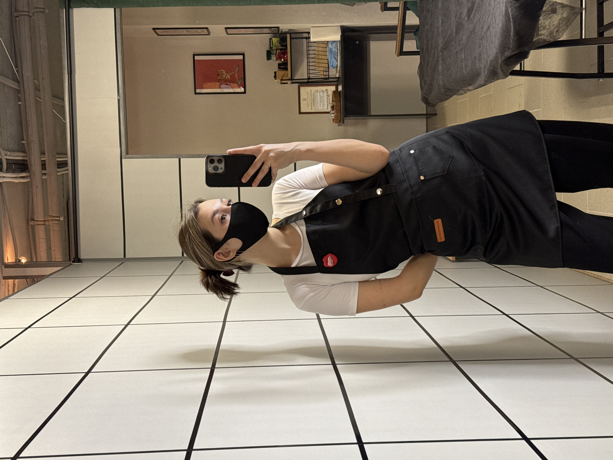 The rotated image captures a person taking a selfie in what appears to be a mirror, from the chest up. The person is wearing a black face mask, a white short-sleeved t-shirt, and a black apron. Their light brown hair is tied back in a low ponytail. They hold a black smartphone, likely an iPhone, with their right hand, angled towards themselves to capture the reflection. A round red pin with a white design is visible on the left strap of the apron. The apron also features a small reddish-brown label on its side, bearing the faint text "MADE IN BRAZIL."
The setting is an indoor space, possibly a backroom or staff area of a commercial establishment. Reflected in the mirror behind the person are several elements: a light-colored wall adorned with framed pictures, one clearly showing a tiger and another appearing to be a certificate with illegible text. A dark fabric item is draped over a structure to the right of the reflection, and a shelving unit with various items can also be seen. The floor is composed of large white tiles with contrasting dark grout lines, forming a grid pattern. To the far left of the image, industrial-style pipes, wires, and part of a light fixture are visible. The lighting is bright and artificial. The person's expression is obscured by the mask and phone, but they appear focused on the act of taking the photograph.