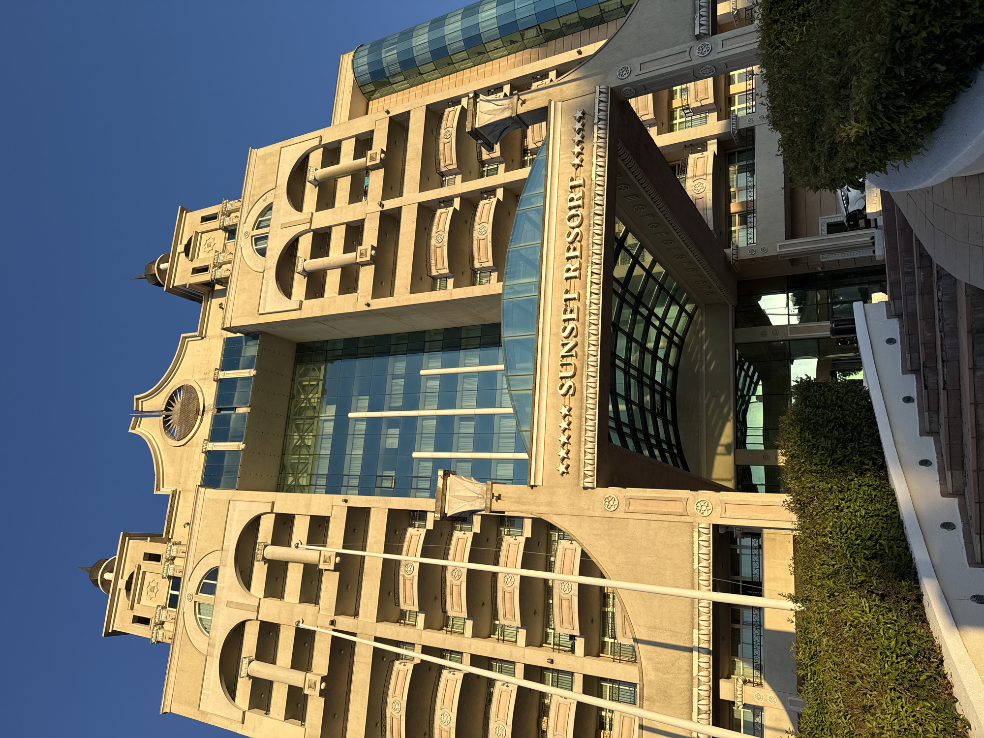 The media file is a photograph of the Sunset Resort in Pomorie, Bulgaria, taken from an upward angle against a clear blue sky. The building's architecture is elaborate, featuring cream-colored facades with arched windows, balconies with ornate detailing, and decorative columns. A prominent glass structure with a curved roof juts out from the building, reflecting the sky and surrounding elements. The name "SUNSET RESORT" is visible in raised lettering on the side of the building, along with decorative stars. Lush green bushes are visible in the foreground, cascading down a terraced area with steps. The sunlight suggests it is either early morning or late afternoon, casting long shadows and highlighting the architectural details. There are no people or animals visible in the image. The scene evokes a sense of grandeur and tranquility.