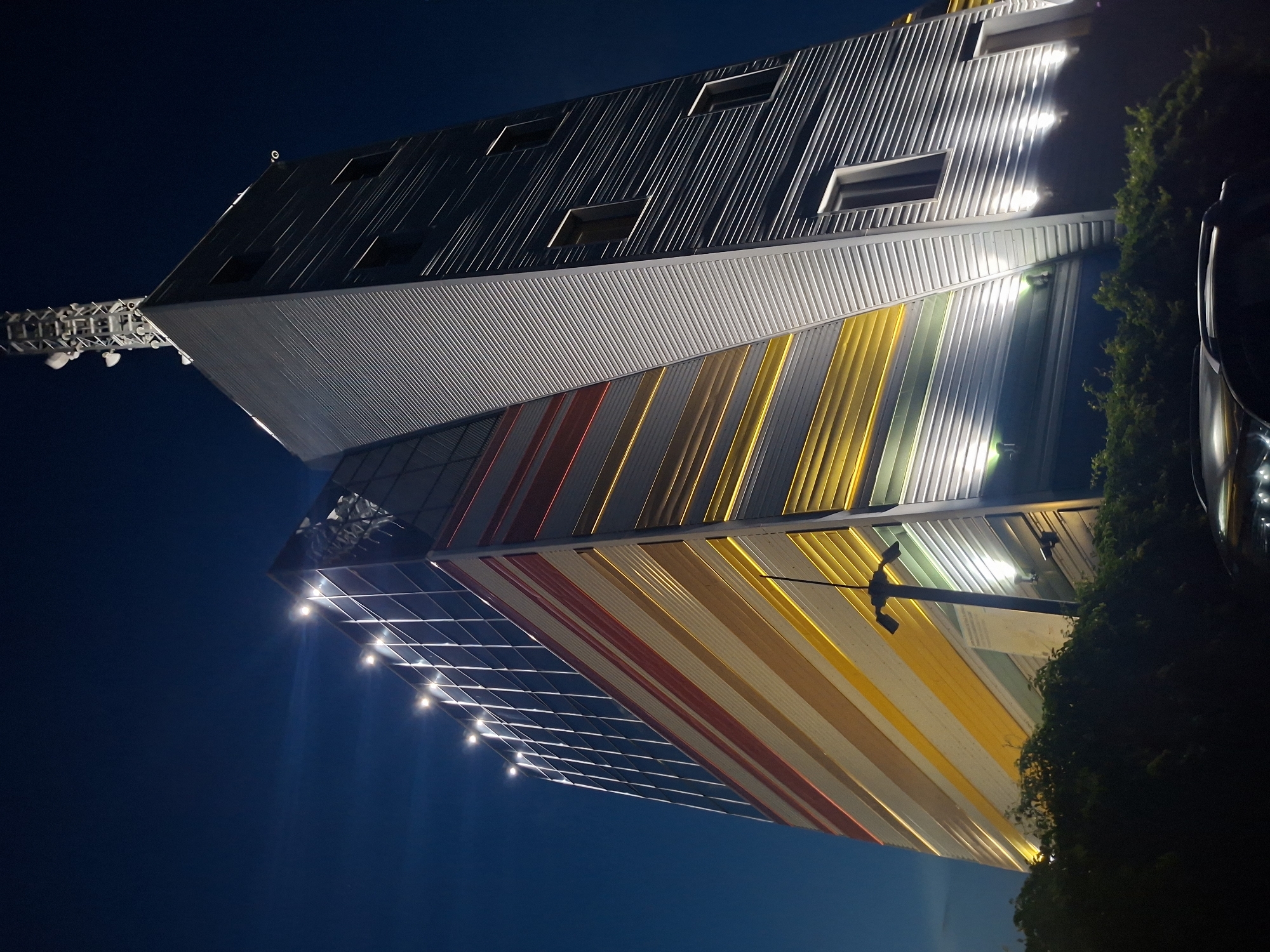 The image shows a tall, modern building at night, located in the city of Varna, Bulgaria. The building has a distinctive facade with horizontal bands of color, including red, yellow, and white, set against a darker metallic material. The upper section of the building features a large communication tower with multiple antennas. The building is illuminated by external lights, casting a glow on its surfaces and the surrounding dark sky. In the foreground, the tops of some lush green trees and the roof of a car are visible, suggesting the building is in an urban or semi-urban setting. The overall impression is one of a contemporary architectural structure illuminated for nighttime visibility.