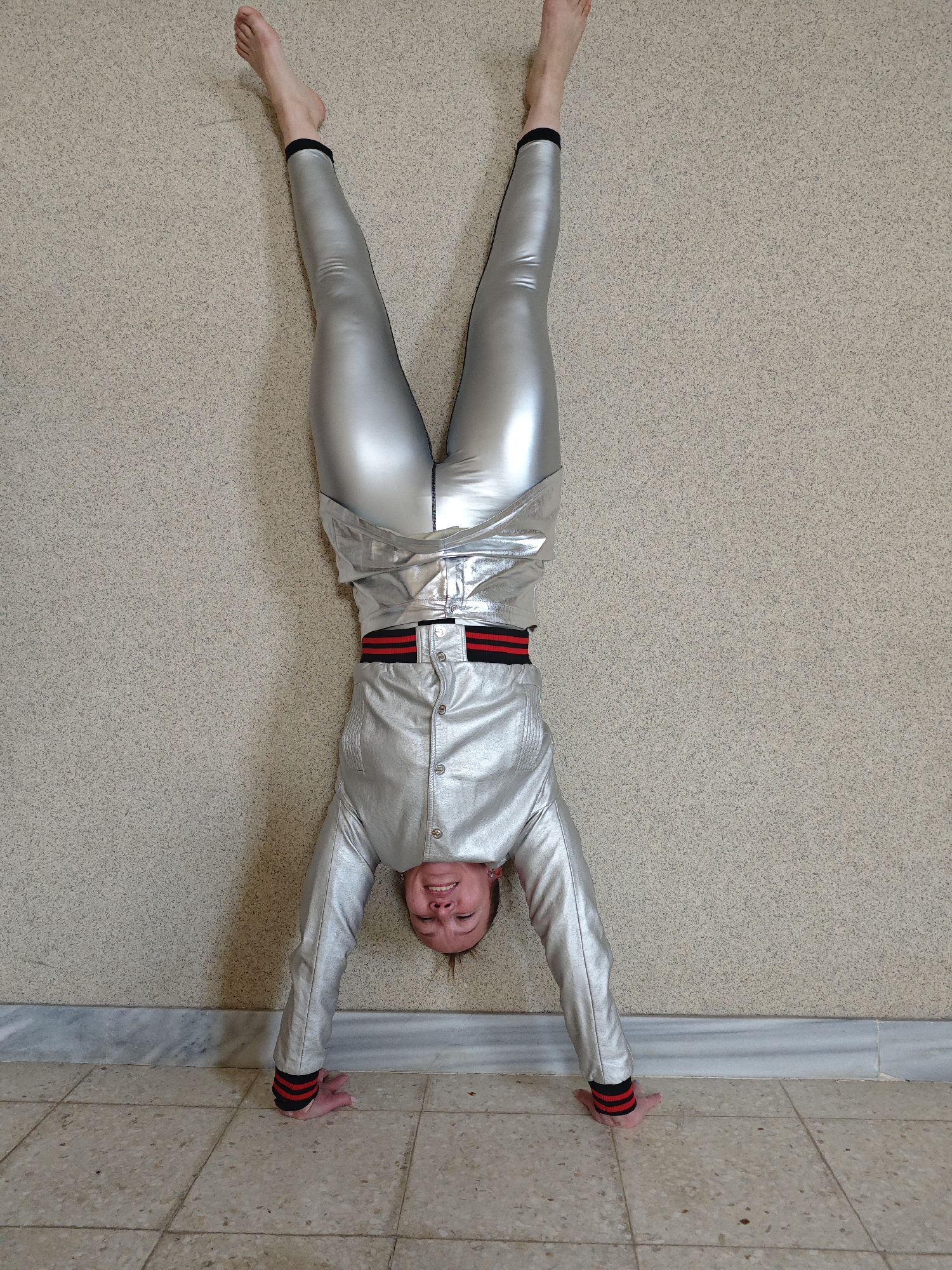 A person wearing silver leggings and a silver jacket with red and black trim is performing a handstand against a textured beige wall. The person is barefoot, and their head is near the beige tile floor. The location is not discernible from the image.