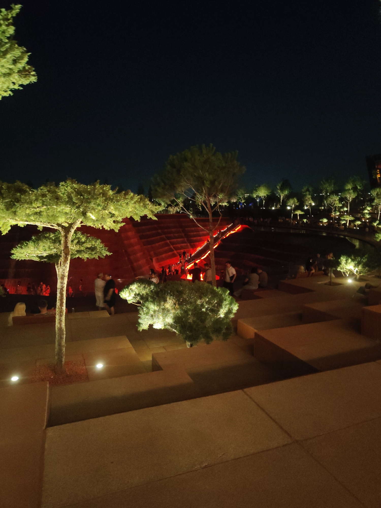 The image depicts an outdoor amphitheater at night, likely part of a park or recreational area. The scene is illuminated by artificial lights, casting a warm glow on the tiered seating, walkways, and surrounding vegetation. In the background, a large, red-lit structure suggests a stage or a prominent architectural feature of the amphitheater. Several people are visible, scattered throughout the seating areas and on the walkways, some appearing to be engaged in conversation or observation. The foreground features trees and shrubs, also lit from below, creating an atmospheric ambiance. The overall impression is one of a lively gathering place, possibly for an event or performance, within a modern landscaped environment. The location is identified as the "Memory of Krasnodar" park in Krasnodar, Russia.